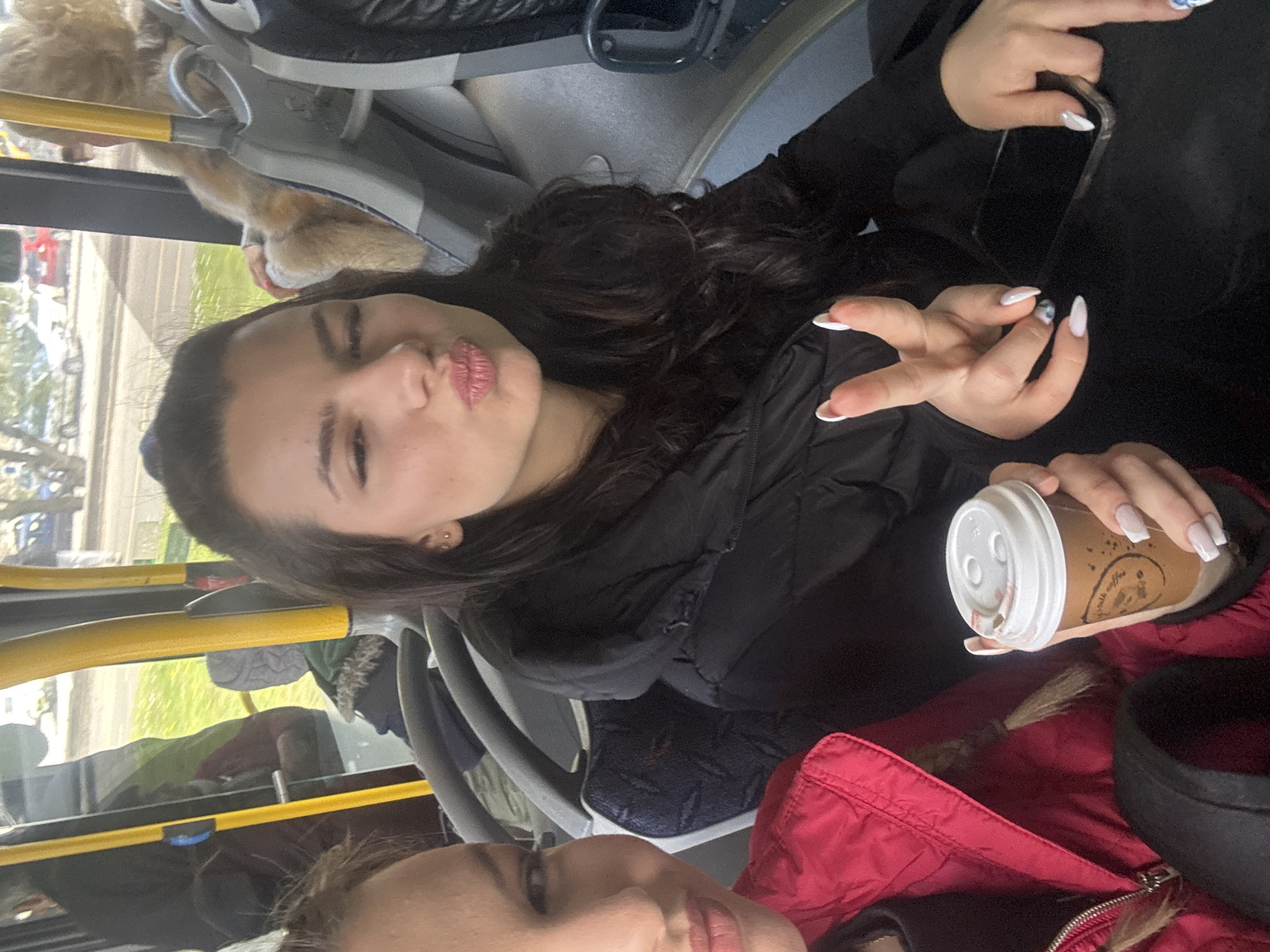 A photo of two young women on a bus, likely taken by one of them. One woman is making a kissy face at the camera, while the other is partially visible at the bottom of the frame, also making a kissy face. The first woman is holding a coffee cup and her cell phone and is wearing a black jacket. The second woman is wearing a red jacket. The interior of the bus is visible, with yellow rails and seats. The background shows an urban scene, which could possibly be Burgas, Bulgaria.