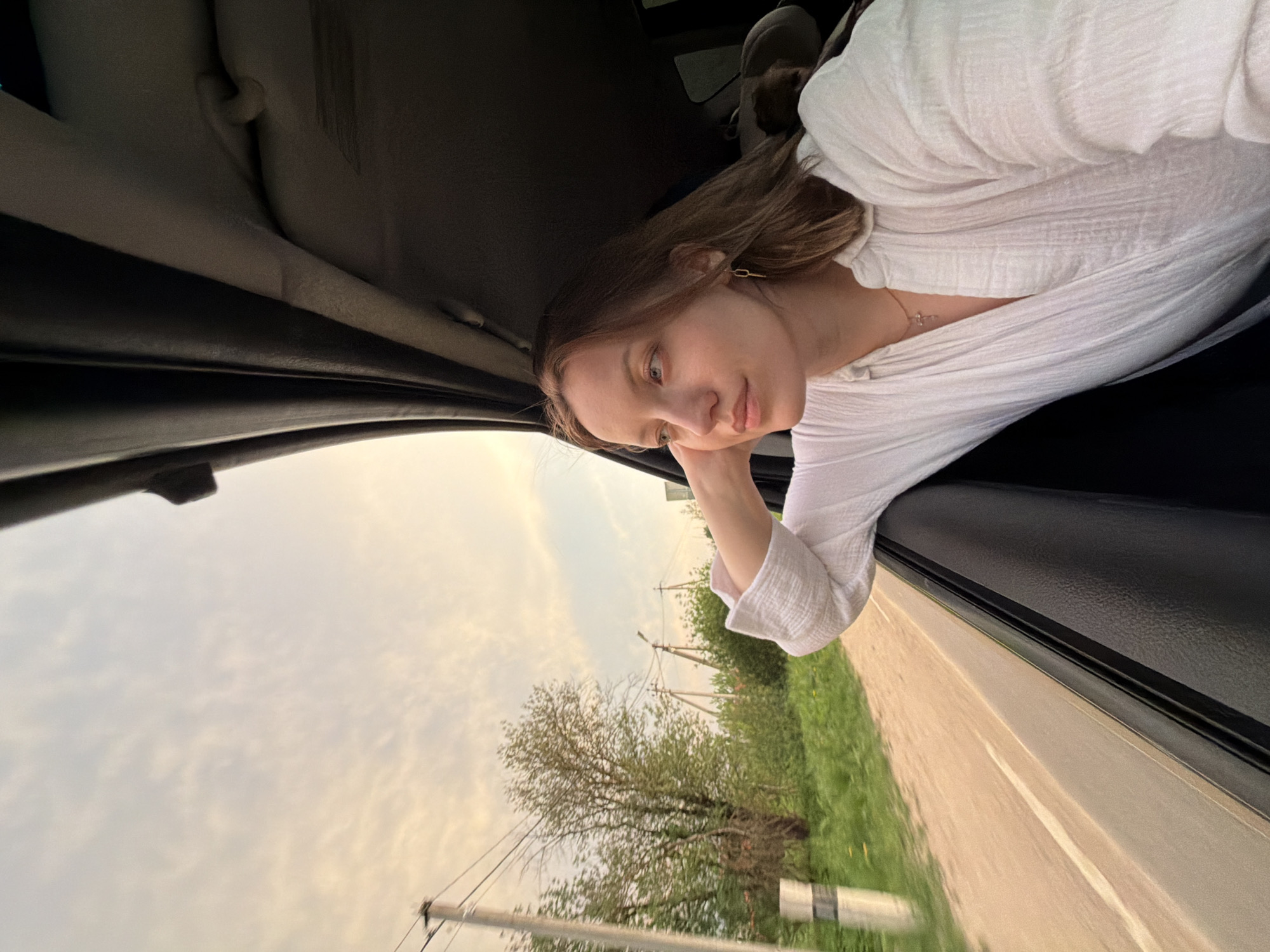 A woman is sitting in a car, leaning her head on her hand and looking out the window. The scenery outside the window shows a rural road, green grass, trees, and power lines. The sky is cloudy. The location is Reshetnikovo, Russia.