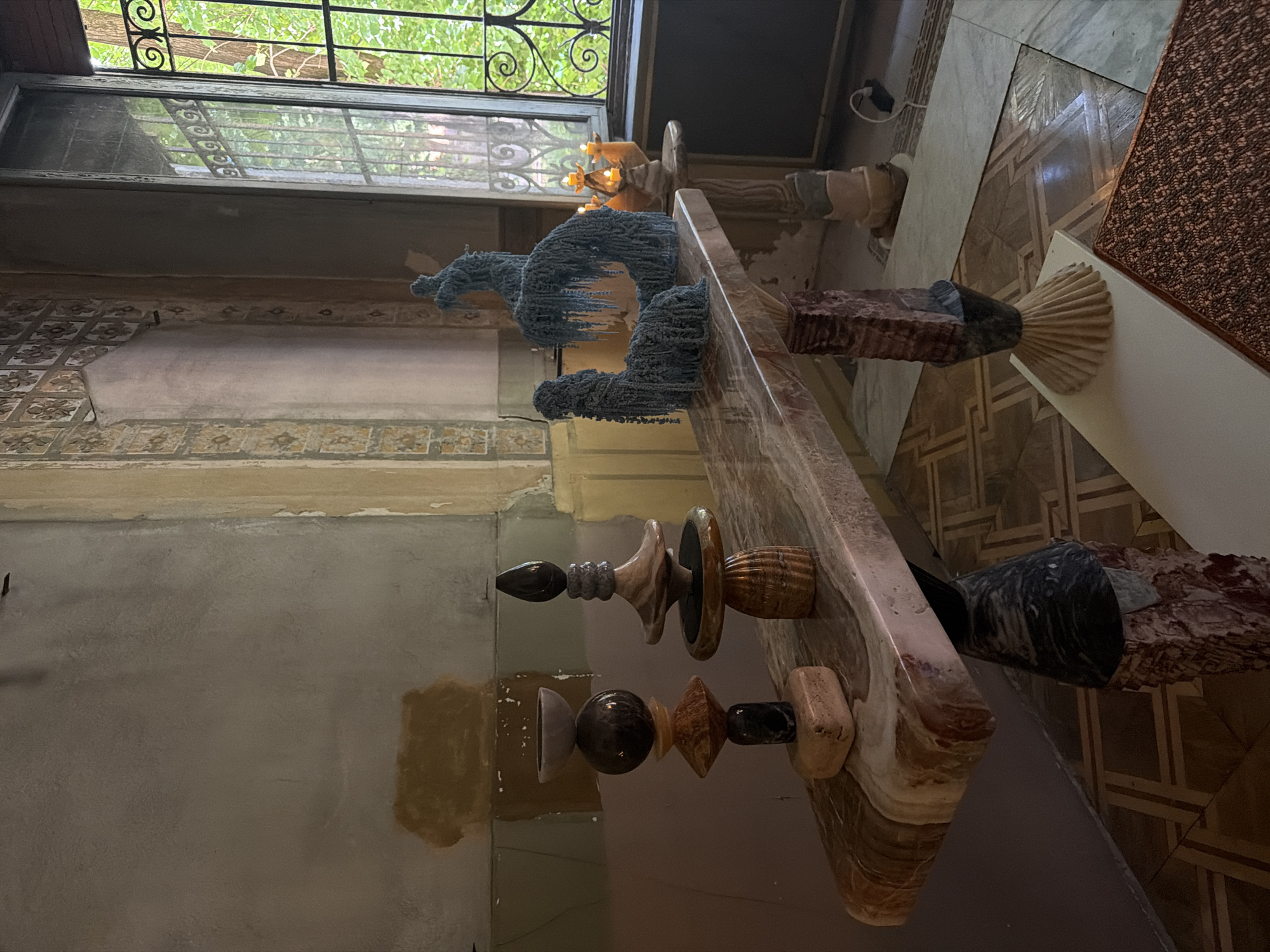 The image depicts an interior in Milan, Italy, featuring a distinctive marble table displaying several objects of varying materials and design. A window is visible in the background.
Objects on the table include abstract sculpted forms appearing to be constructed from textured blue material. There are also several other sculptures made from wood and marble in different designs.
The table is marble with prominent veining patterns. The floor is patterned. The surrounding wall is partly covered with decorative patterns and partially faded paint.