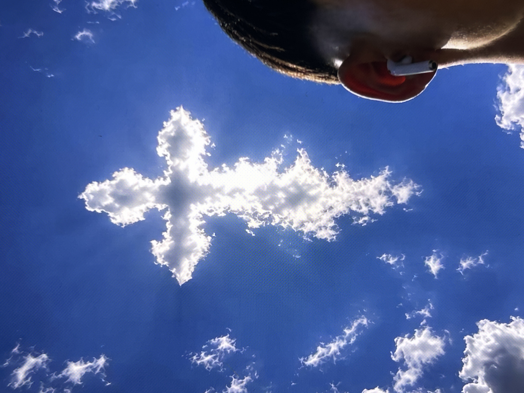 The image shows a person's head and ear, with a white earbud in the ear, positioned in the top right corner. Below, a cloud formation in the shape of a cross dominates the blue sky. Smaller cloud formations are scattered throughout the rest of the sky. There is no information present in the image to indicate that the photograph was taken in Zetiovo, Bulgaria.