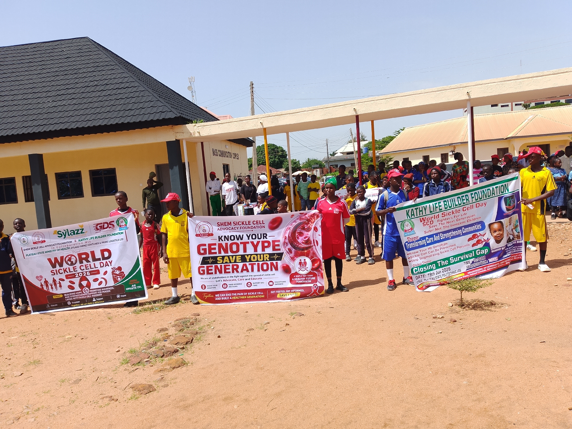 In Jos, Nigeria, on a sunny day, a group of people, many of them young individuals, are gathered outside a light yellow building with a dark grey tiled roof. They are holding up banners for a sickle cell awareness event. The building appears to be the "MASS COMMUNICATION COMPLEX."
The main focus is on the banners, which are colorful and informative. One banner, held by a young man in a red cap and yellow shirt, reads "KATHY LIFE BUILDER FOUNDATION" and "World Sickle Cell Day - Save The Next Generation." Another banner, held by a group of people in the center, displays "SHEM SICKLE CELL ADVOCACY FOUNDATION" and "KNOW YOUR GENOTYPE - SAVE YOUR GENERATION." A third banner, held by individuals on the left, states "SyLazz" and "WORLD SICKLE CELL DAY - 2026." These banners also contain information about themes such as "Transforming Care And Strengthening Communities" and "Building Hope, Promoting Awareness, Together, we can make a difference."
The people are diverse in age and attire. Some are wearing athletic wear, while others are in more casual clothing. The environment is dusty with scattered stones and a small tree. The sky is clear and blue. The event appears to be an awareness walk or rally, with participants posing for a photograph.