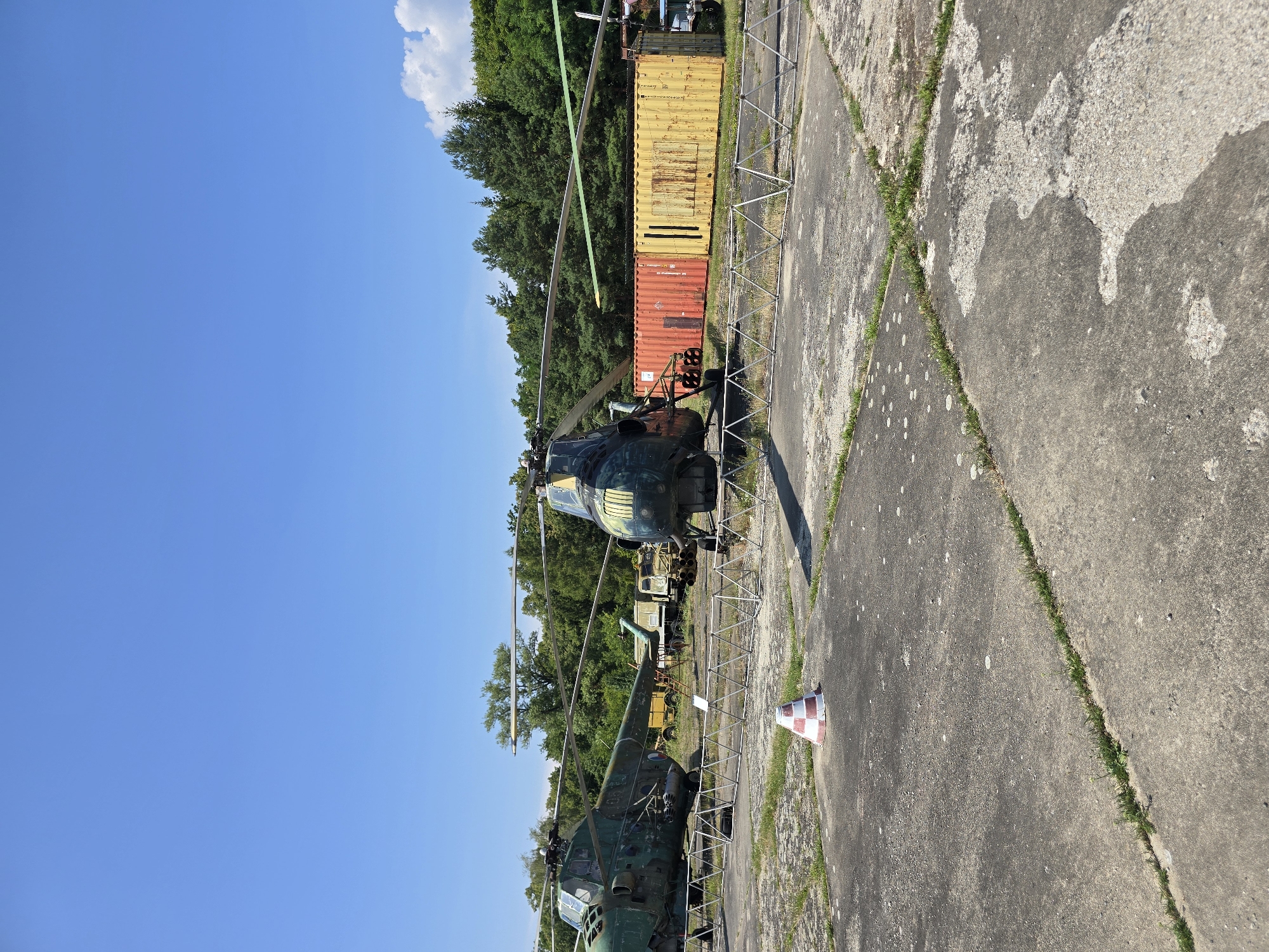 Two weathered helicopters are parked on a concrete tarmac under a clear blue sky. The helicopter on the left is predominantly green with patches of rust, while the helicopter in the center is a darker color with yellow markings on its front. Both helicopters have visible rotor blades. In the background, a line of trees forms the horizon. Between the helicopters and the trees, two shipping containers, one orange and one yellow, are visible, along with other unidentifiable vehicles. A low metal fence runs across the foreground, separating the tarmac. A red and white striped traffic cone stands on the concrete in the mid-ground. The scene appears to be an outdoor storage or display area for disused aircraft.