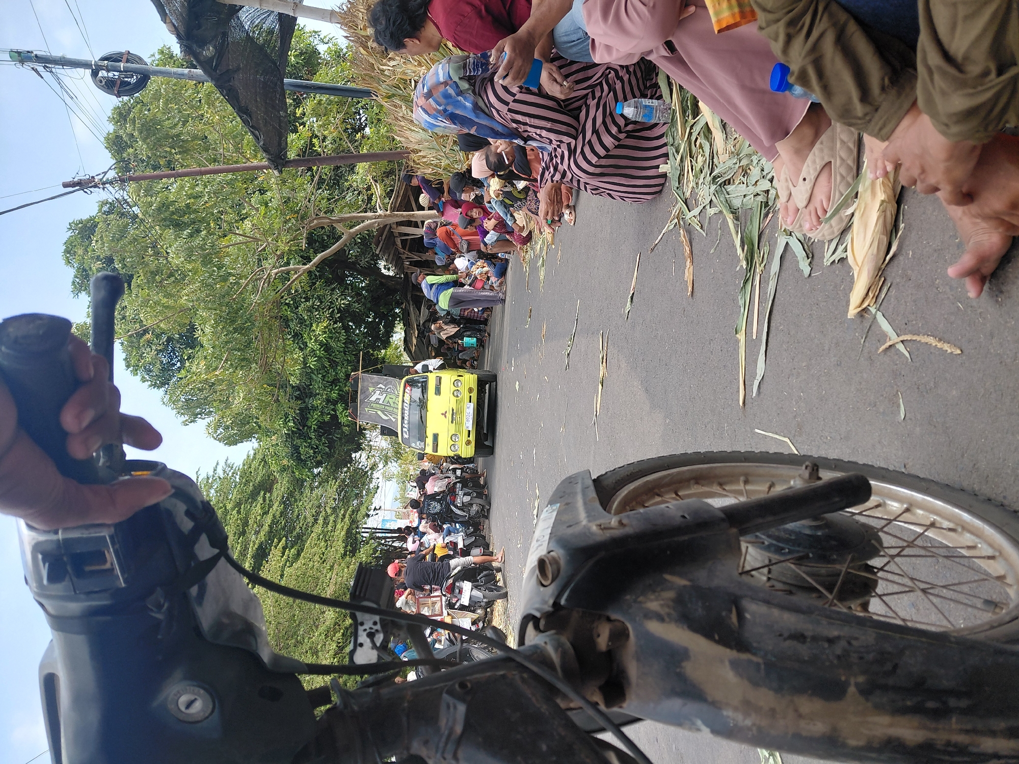 The image shows a scene from a roadside in Bimo, Indonesia, possibly during a local event or gathering. In the foreground, the handlebars and front wheel of a motorcycle are visible, suggesting the photographer is on a motorcycle. The road is lined with trees and a blue sky is overhead. A yellow truck with a banner is parked on the road. Several people are gathered along the side of the road, some sitting on the asphalt and others standing near motorcycles. There are also piles of dried corn stalks and husks on the ground, indicating a harvest or a related cultural activity. The overall atmosphere appears to be one of community gathering or a local festival.