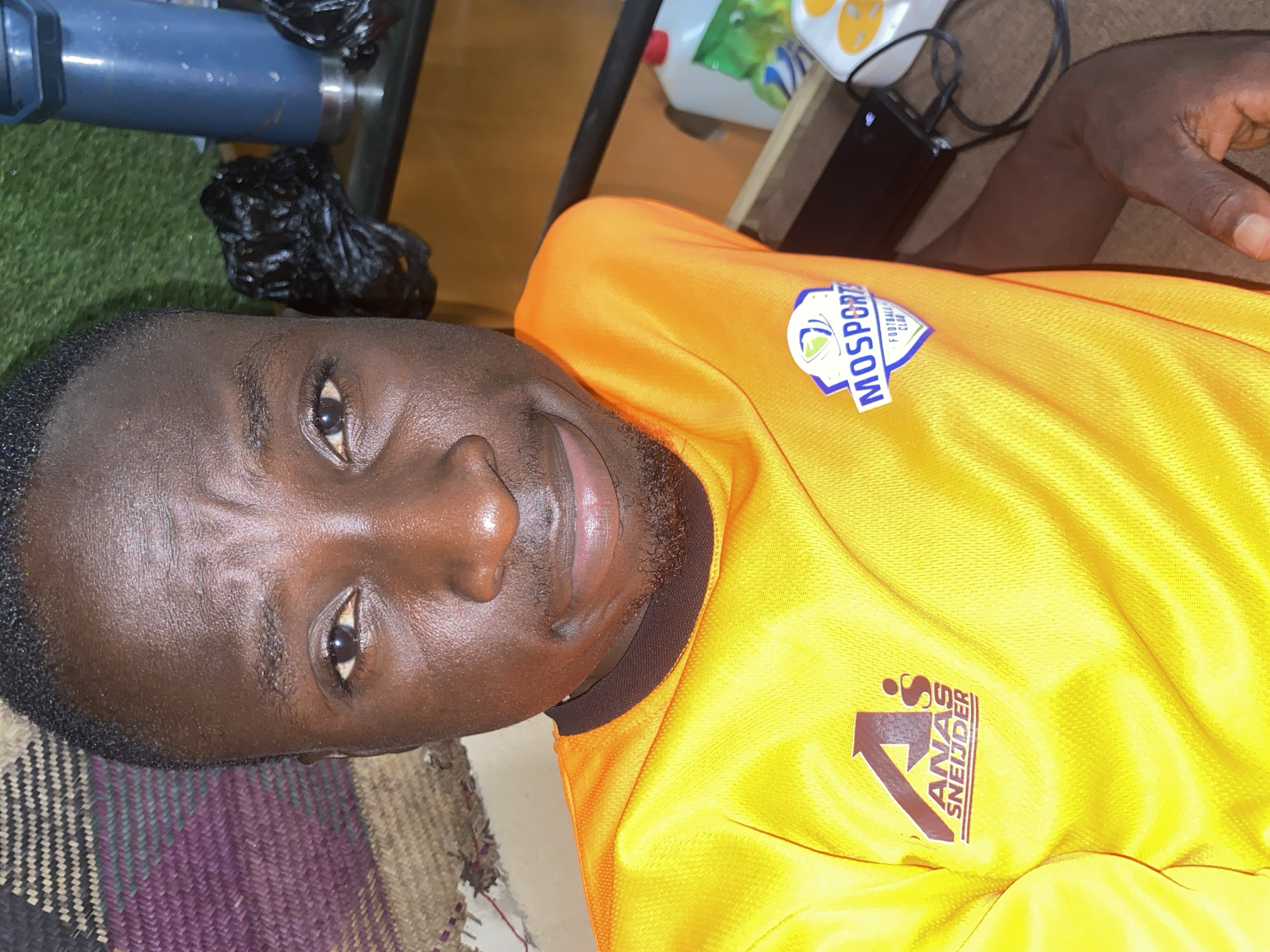 In Barden Yana, Nigeria, a young man, likely in his late teens or early twenties, is captured in a close-up, slightly upward-angled selfie. He is wearing a bright orange t-shirt with two prominent logos. The logo on the left features a stylized 'A' with an arrow and the text "ANAS SNEIJDER" below it. The logo on the right is a shield shape with "MOSPORT FOOTBALL CLUB" inscribed within it. His skin is a deep brown, and he has short, dark hair. His eyes are dark and open, looking towards the camera, with a subtle smile on his lips. The background is a mix of textures and colors. To the left, a woven mat with shades of purple and brown is visible. To the right, a blue insulated water bottle and black plastic bags are placed on a reflective surface, possibly a shelf, with a green, grass-like material behind them. The lighting suggests an indoor setting, possibly at night or in a dimly lit room.