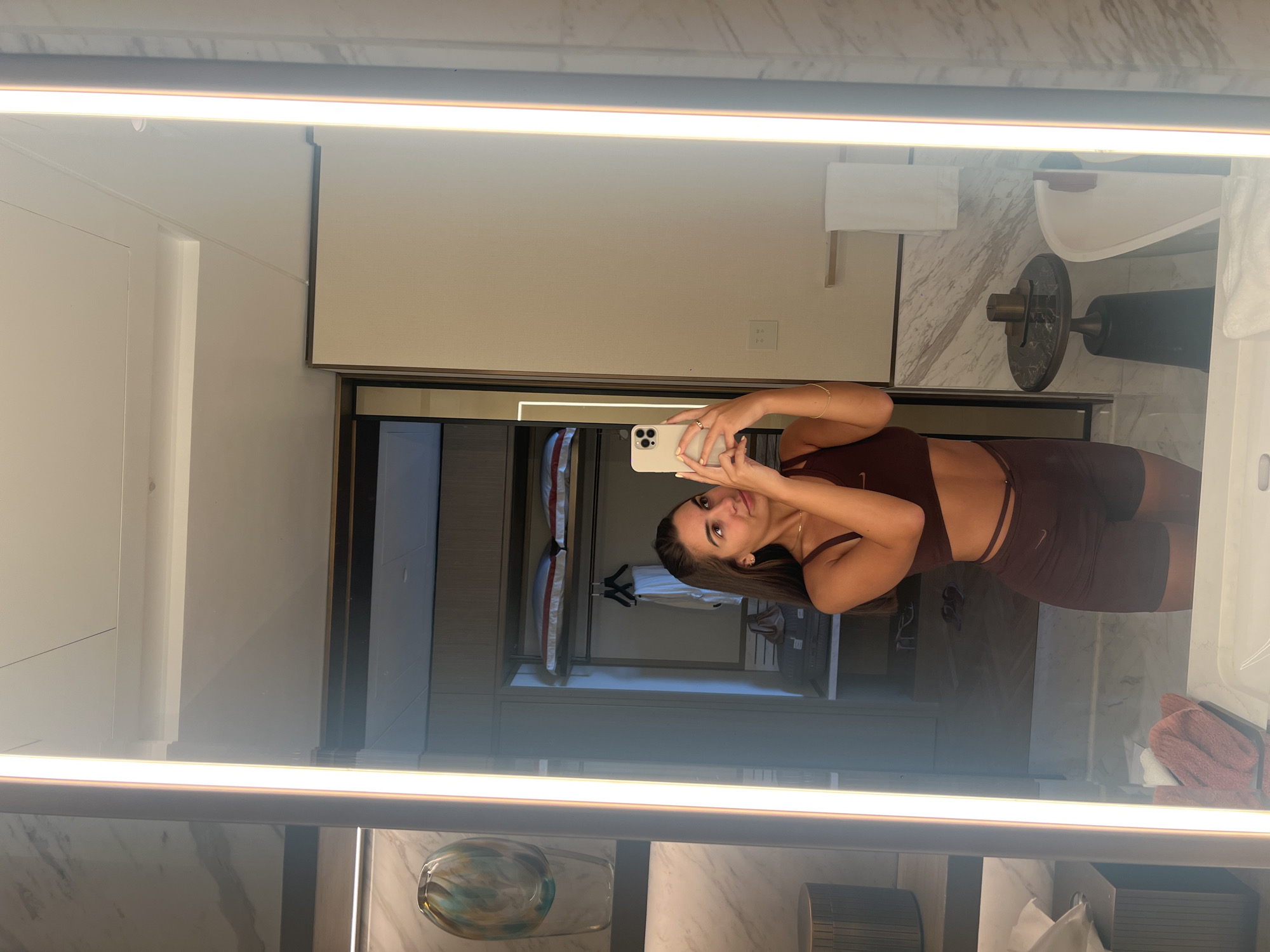 A woman is taking a selfie in a bathroom mirror, which reflects the bathroom behind her. The location is likely in Dubai, United Arab Emirates, based on the style of the room and the presumed wealth suggested by the scene. The bathroom features a modern design with a light-up mirror and marble-style surfaces. A bathtub and a towel rack are visible in the background, and through an open doorway, one can see closet shelves with clothing. The woman is wearing a matching crop top and shorts set and is holding her phone up to take the photo.