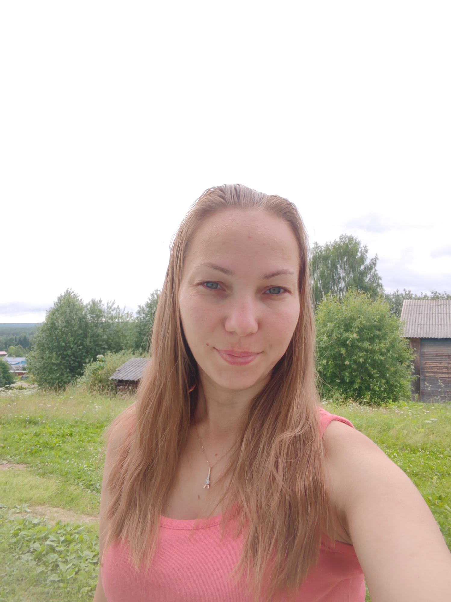 This is a selfie taken outdoors in a rural setting, possibly in or near the city of Chukhlem, Russia. The subject is a woman with long, light brown hair, wearing a pink tank top and a necklace. She is positioned in the foreground, with a slightly smiling expression. Behind her, the background consists of lush green vegetation, including trees and tall grass, with a few small wooden structures visible. The sky is overcast.