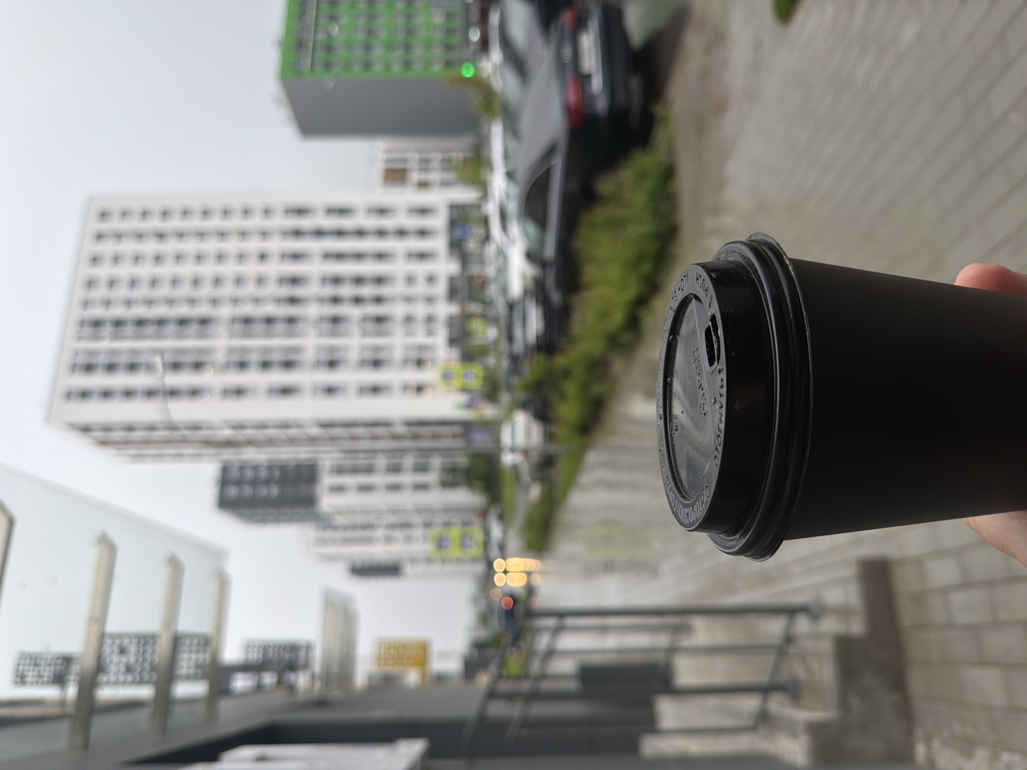 A first-person perspective shot captures a black coffee cup held by a person in the foreground, with a city street scene blurred in the background. The street is wet, indicating recent rain, with reflections of lights from vehicles visible on the pavement. In the mid-ground, several cars are parked and on the road. The background is dominated by tall, modern buildings, some white with a grid-like pattern of windows, and one building colored bright green. The sky is overcast and gray.