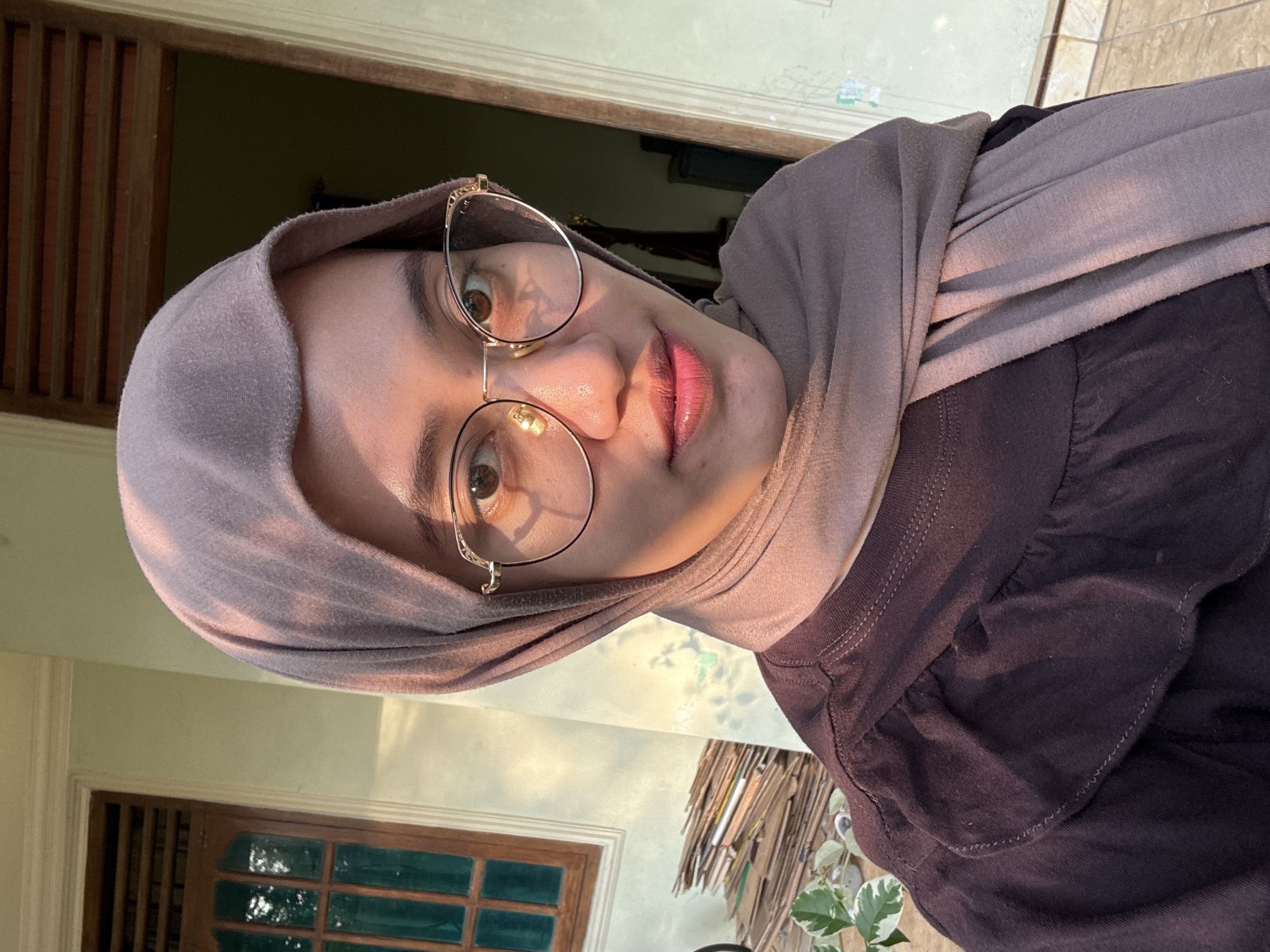 A young Indonesian woman, wearing a brown hijab and glasses, poses for a selfie outdoors in Gadingrejo, Indonesia. She is looking directly at the camera with a slight smile. The background shows a light green wall, a wooden window with green panes, and a doorway. To the left of the woman, a stack of cardboard boxes is visible, along with some greenery. The setting appears to be residential, possibly a porch or courtyard, with the warm light of the sun casting shadows.