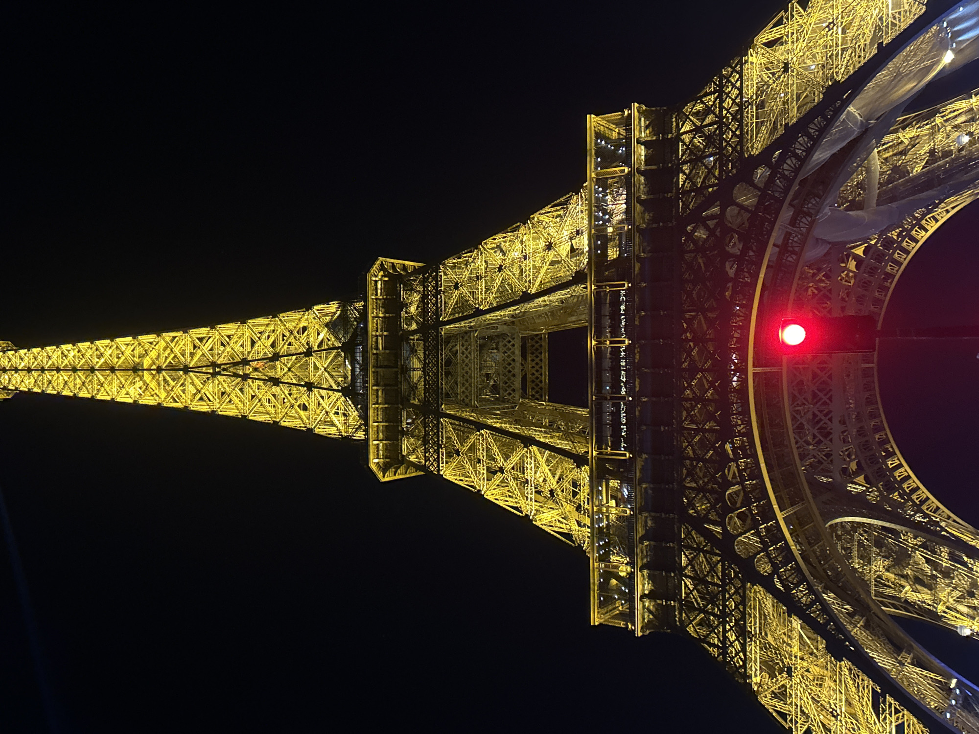 This image depicts the Eiffel Tower at night in Paris, France. The tower is illuminated with golden lights and has a red light visible on one of its levels. The background is a dark, almost black, night sky. The camera angle is looking upwards, emphasizing the tower's height and structure.