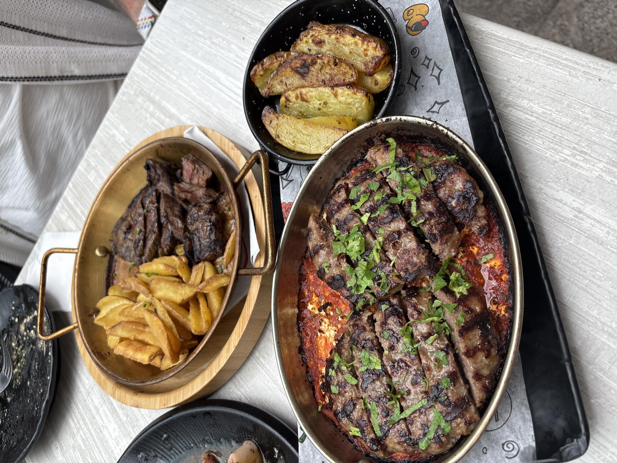 The image captures a close-up view of a table setting at a restaurant, likely in Burgas, Bulgaria, given the context. The focus is on a meal featuring grilled meat and potatoes.
In the foreground, a metallic serving dish holds two large, grilled patties, sliced and topped with fresh chopped parsley. These patties are situated atop a bed of what appears to be a red sauce, possibly tomato-based, with some visible chunks of cheese or feta.
To the right, a small black bowl contains several wedges of golden-brown roasted potatoes, some showing slightly darker, caramelized edges.
In the background, a larger round wooden tray with metallic handles contains more grilled meat, sliced, alongside a generous portion of wedge-cut fries. The serving dishes are placed on a table with a light-colored, textured surface, possibly wood or a wood-like laminate. A dark serving tray or placemat is visible underneath the main dishes. A partially visible black plate with remnants of food and a fork is in the upper left corner, suggesting other dishes have been served or are being consumed. The overall lighting suggests an outdoor or well-lit indoor environment, with a hint of a person's white clothing visible at the top right of the frame.