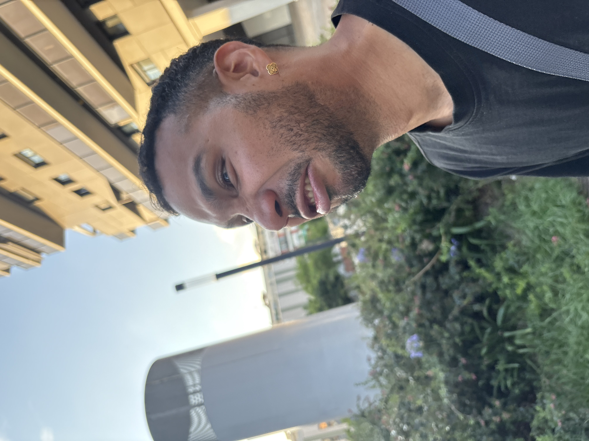 The image features a man in the foreground, captured from a slightly low angle with his head tilted. He has dark, curly hair, a beard, and a mustache. A gold-colored earring, shaped like a four-leaf clover, adorns his left ear. He is wearing a dark t-shirt, with a grey strap visible over his right shoulder, suggesting he might be carrying a bag. His mouth is slightly open, revealing his tongue, and his eyes are looking towards the viewer, conveying a casual or possibly playful expression.
The background depicts an outdoor urban environment, consistent with the provided context of Barcelona, Spain. To the left, modern, multi-story buildings with numerous windows are visible under a bright, clear sky. In the mid-ground, a tall, light-colored cylindrical structure and a street lamp pole are present. To the right of the man, a dense arrangement of green foliage, including bushes and some small purple flowers, forms a natural backdrop. The lighting suggests it is daytime, with bright, diffused light indicating either morning or late afternoon. There is no visible text in the image.