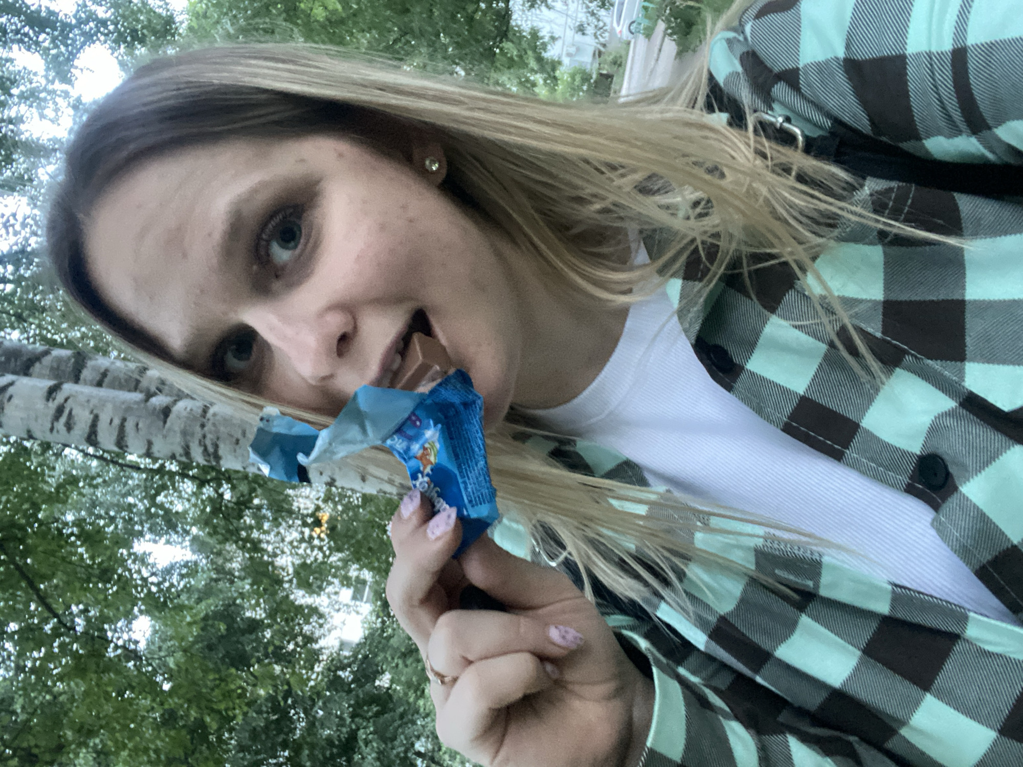 A light-skinned woman is captured in a selfie, holding a blue candy wrapper in her left hand and with a piece of chocolate in her mouth. She is wearing a white shirt under a green and black plaid jacket. Her nails are painted with light pink polish. The background reveals foliage and a birch tree trunk. It's hard to confirm the location with absolute certainty, but based on available visual cues, it could plausibly be the city of Chaikovsky, Russia.