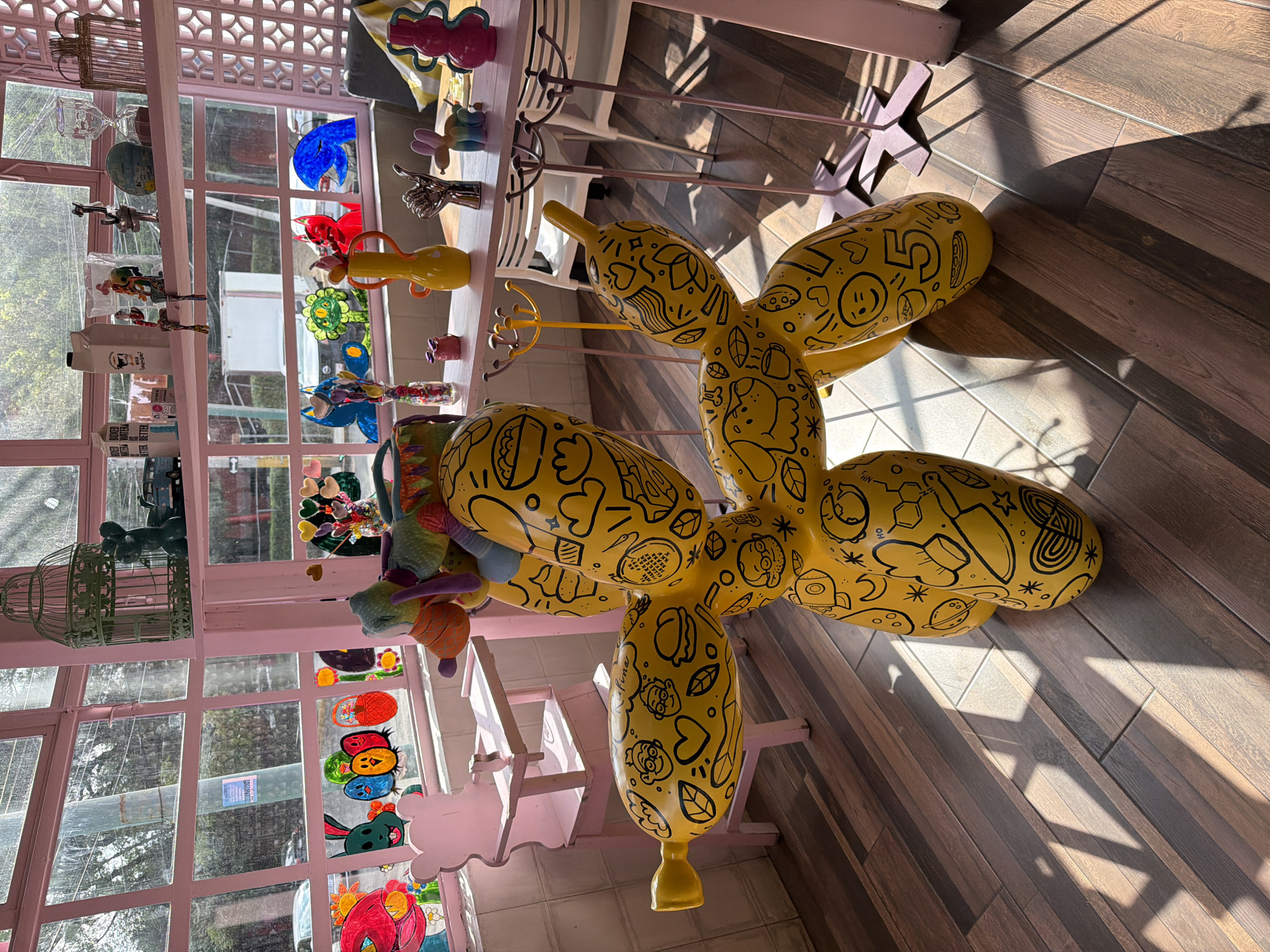 The image shows a yellow balloon dog sculpture with black doodle-like patterns on a light brown, wood-patterned floor. The sculpture is located inside a room with a pink-framed window. The window shows a scene with trees and sky. In the background are display shelves also painted pink with assorted trinkets and colorful glass objects on them. The scene is well-lit, with shadows indicating sunlight entering the room. There are no visible people in the frame. The location appears to be a shop or cafe interior, likely in Puebla, Mexico.