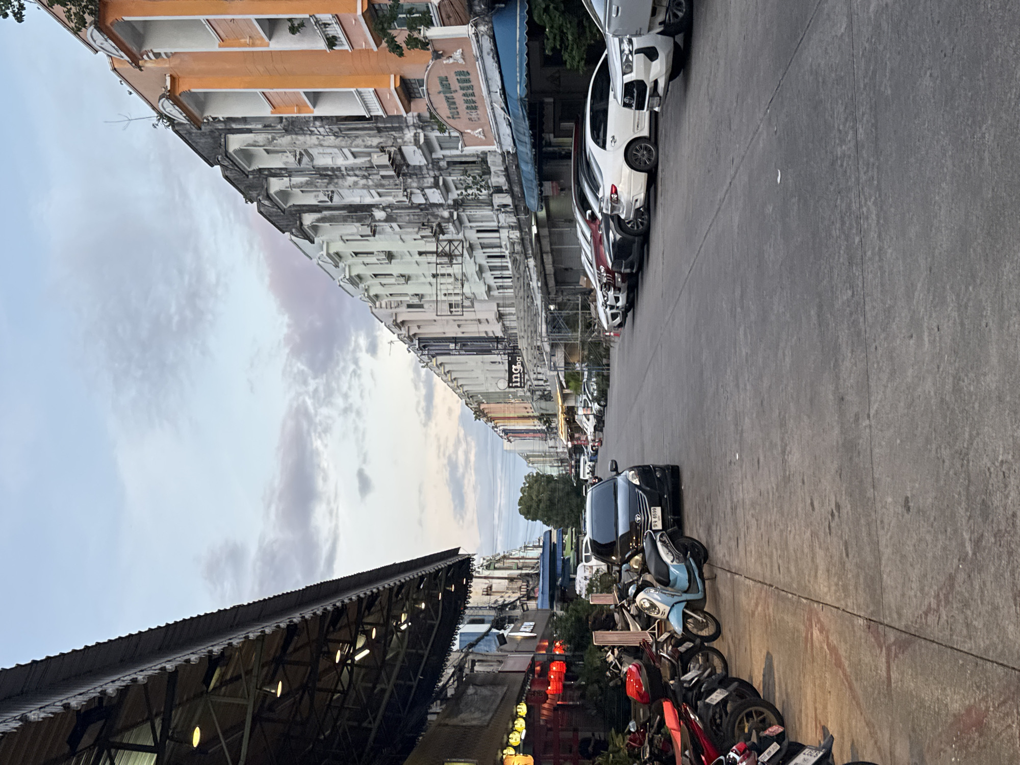 The image captures a street scene in Phuket City Municipality, Thailand, with a row of buildings lining the right side of the road and an awning extending over a sidewalk on the left. The buildings are a mix of architectural styles and colors, with some appearing older and weathered, while others are more modern and brightly painted, such as the orange and cream building at the far right. The sky is overcast with soft clouds.
On the left side, a covered walkway with lights and red lanterns suggests commercial establishments, possibly restaurants or shops. A variety of motorcycles are parked along the sidewalk. A light blue scooter is prominent in the foreground. A dark-colored sedan is parked in the street near the motorcycles.
The street itself is paved and appears to be a two-way road with cars parked along the right side. Several cars are visible, including white sedans and a darker colored SUV. The overall impression is of a moderately busy street in a tropical urban environment, possibly in the late afternoon or early evening given the soft lighting. There are no people prominently featured or engaged in any discernible actions. The scene is static and depicts a typical streetscape.