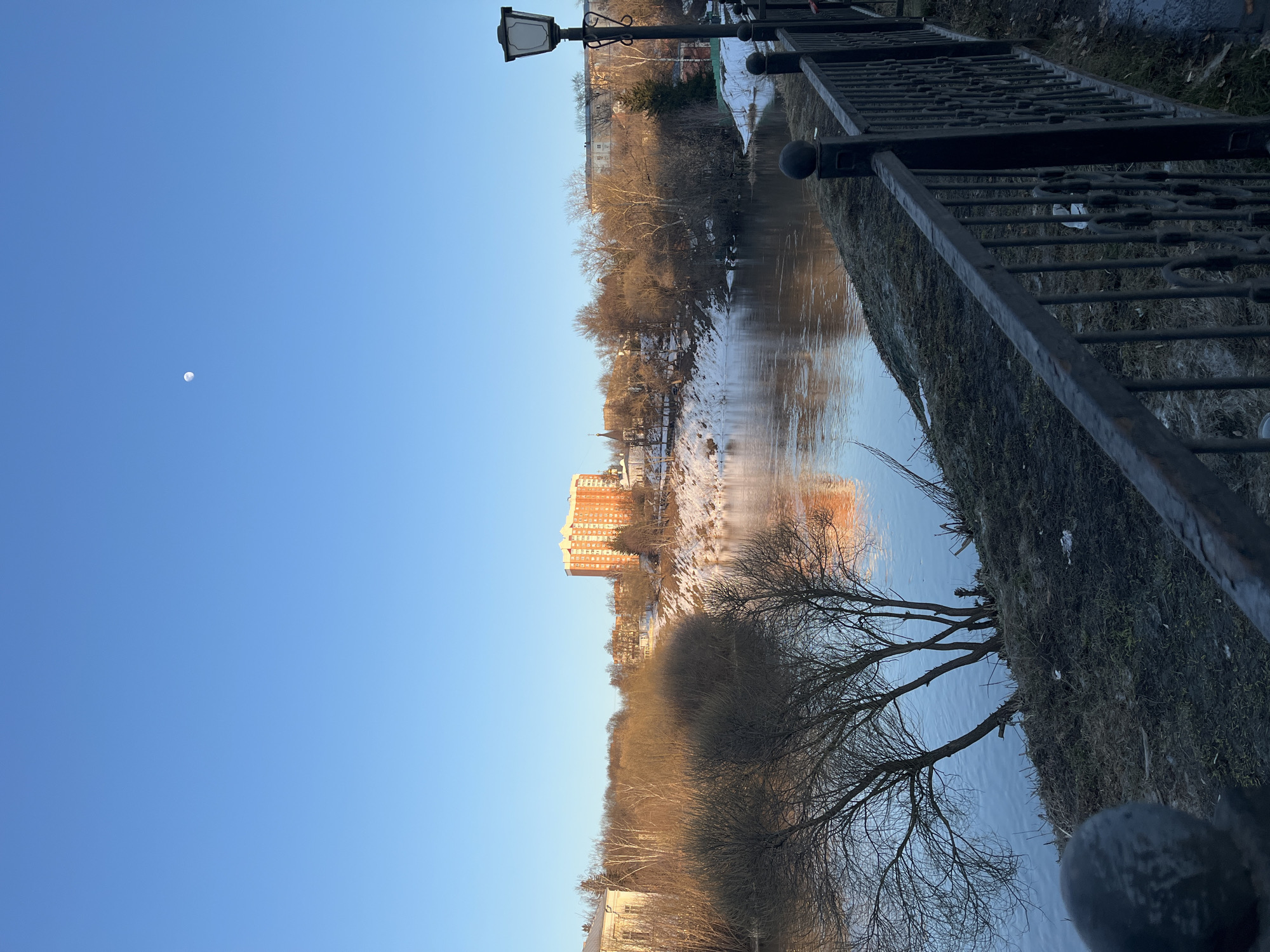 The image shows a view of a river with trees on both banks and a building in the background. The photo was taken in Podolsk, Russia, as indicated by the architectural style of the building. The river reflects the building and the sky. A metal railing is visible in the foreground. The sky is blue, and there is a small moon visible. There is snow on the ground on the riverbanks. A lamppost is positioned near the railing.