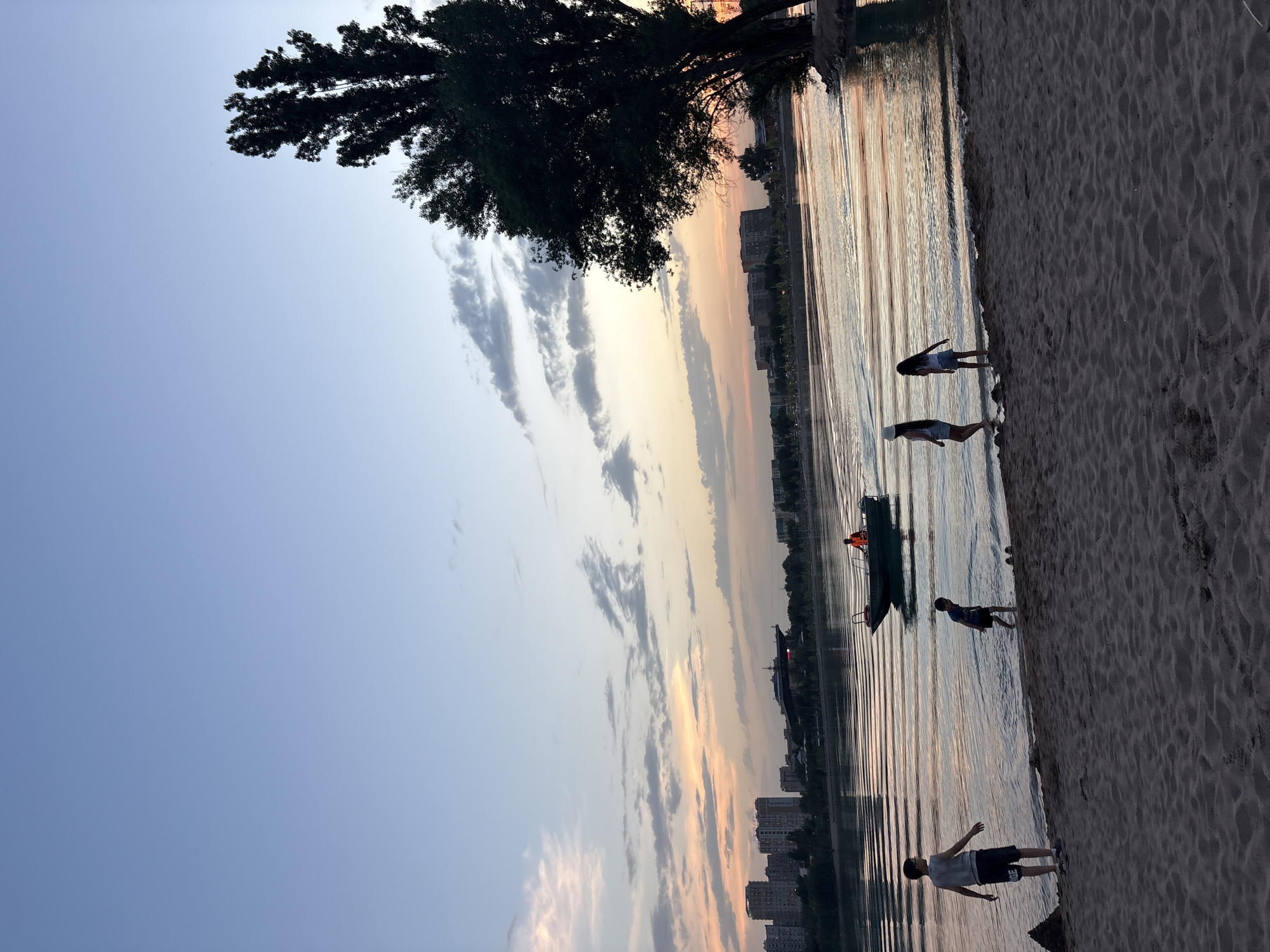The image depicts a sunset scene at the Large Almaty Lake, Kazakhstan. In the foreground, a sandy beach is visible with several children playing near the water's edge. One child, a boy wearing a gray t-shirt and dark shorts, stands on the sand facing the water. Another boy, in dark shorts and a lighter t-shirt, is in the shallow water. Two girls, one in a white hat and shorts and the other in dark shorts, are also near the water. In the middle ground, a small boat with a person wearing an orange vest is moving across the lake. The background features a cityscape with buildings and a bridge structure, all silhouetted against a colorful sunset sky with scattered clouds. A large tree is prominently visible on the right side of the image, its branches silhouetted against the sky. The lake water reflects the colors of the sunset, creating a serene atmosphere.
