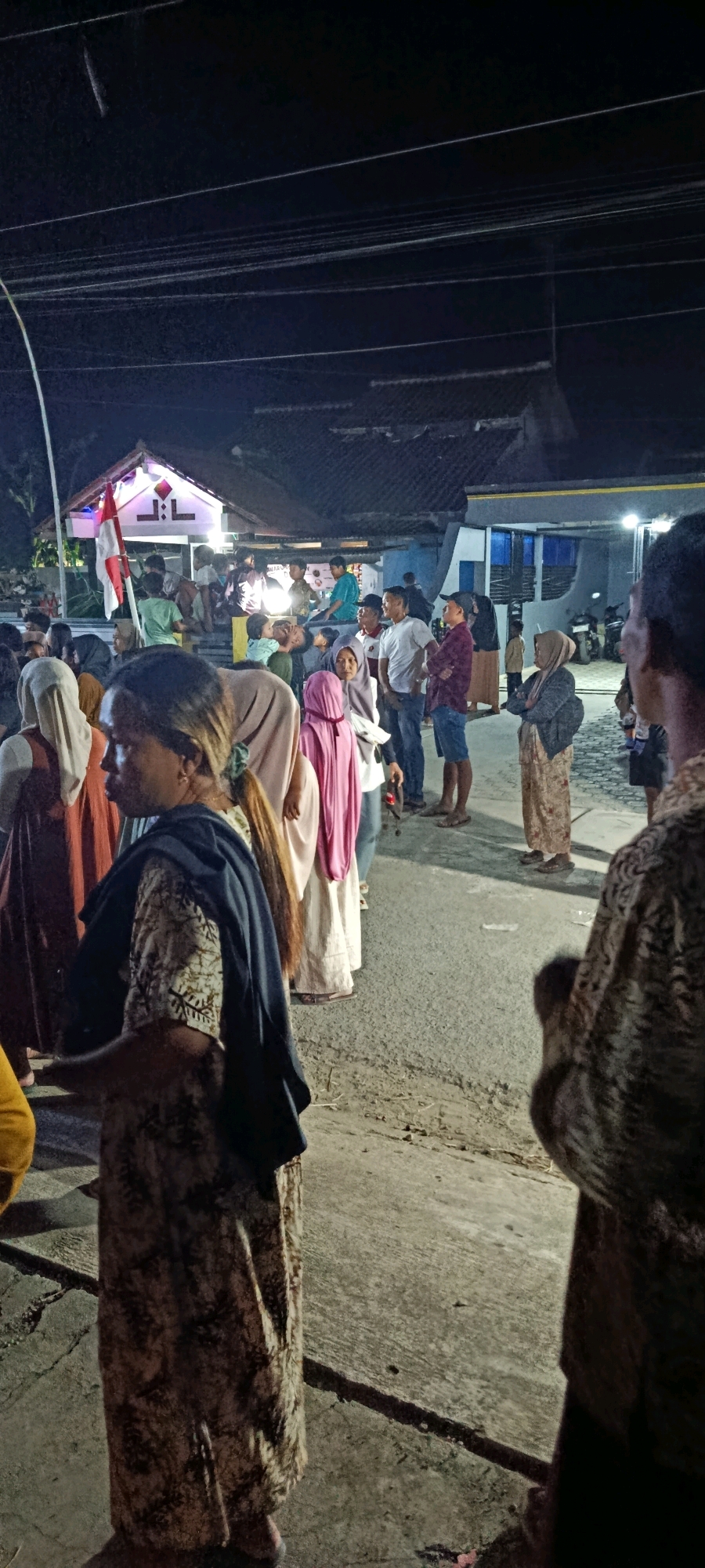 A crowd of people are gathered on a paved street at night. The scene is illuminated by artificial lighting from a building in the background, which has a red and white flag displayed on its left side. Several individuals are wearing headscarves. The people are of various ages and are standing and interacting in small groups. The environment appears to be a residential area, with houses visible behind the crowd. The overall atmosphere suggests a community gathering or event.