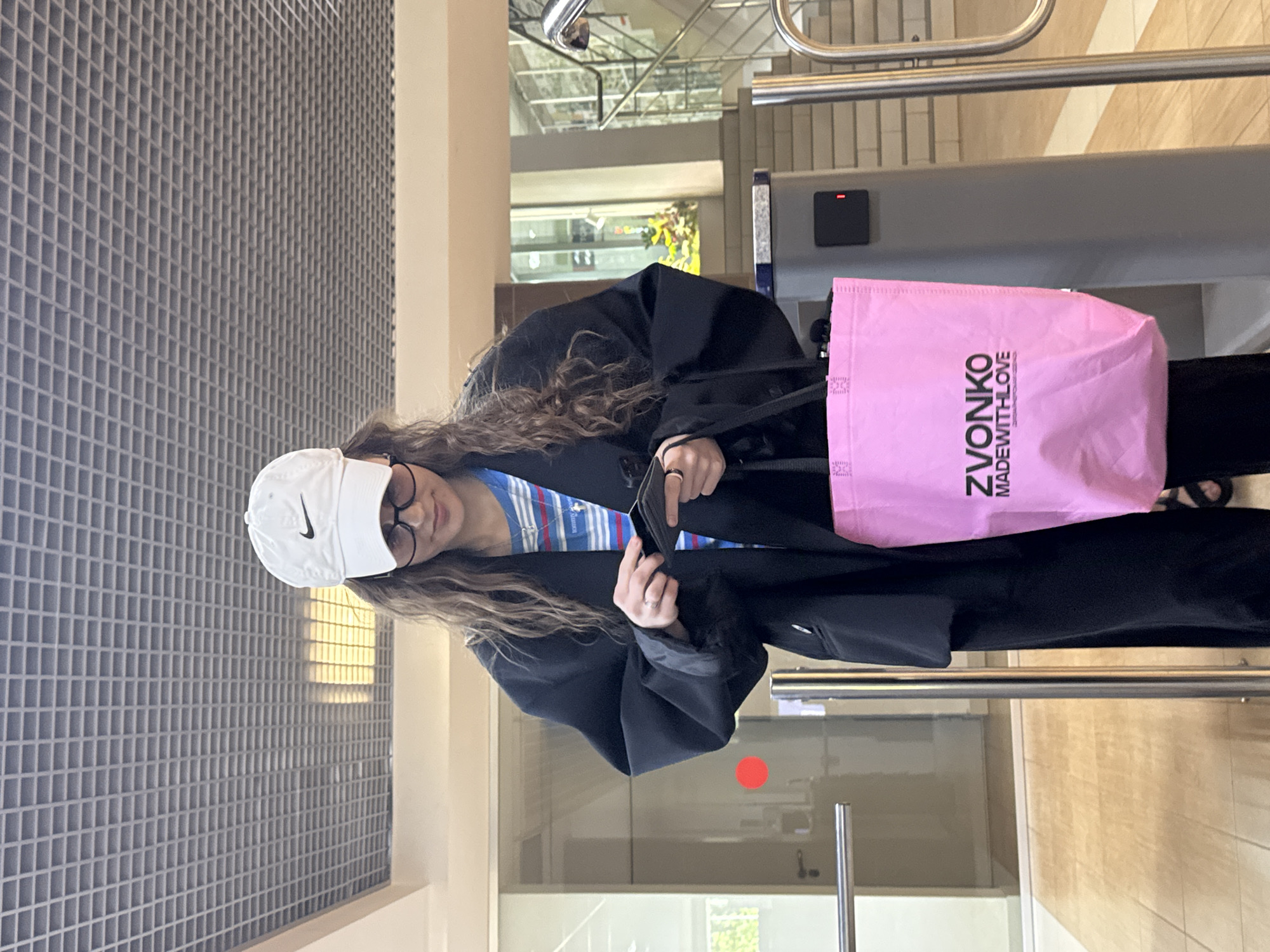 A young woman stands in what appears to be a modern, well-lit indoor space, possibly an office building or public building in Perm, Russia. The woman is wearing a white Nike baseball cap, glasses, a striped shirt, and a long black coat. She holds a black item in her hands, possibly a wallet or phone. A pink shopping bag with the word "ZVONKO" and "MADEWITHLOVE" printed on it hangs from her shoulder. The environment features a grid-patterned wall, a beige pillar, and a background that suggests a doorway or entrance. The floor is tiled in a light beige color. A turnstile is visible in the background. The scene is brightly lit.