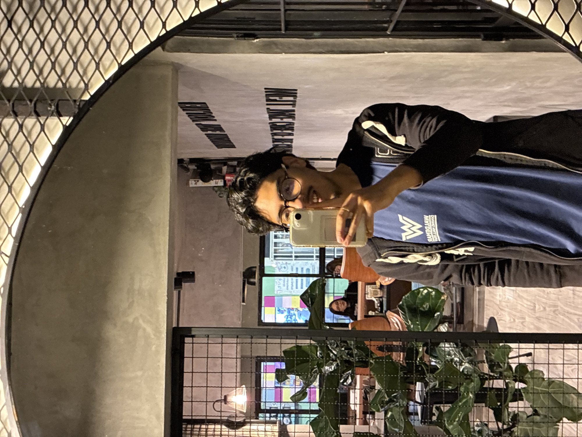 A person is seen in the reflection of a round mirror hanging on a wall indoors. The person is holding a phone and appears to be taking a selfie. The person is wearing glasses, a blue shirt, and a black jacket. The reflection shows a restaurant interior with other people seated at tables and a potted plant in the foreground. The window displays a colorful pattern. Based on the text reflected in the mirror and other details, the location is likely a restaurant in San Andrés Cholula, Mexico.