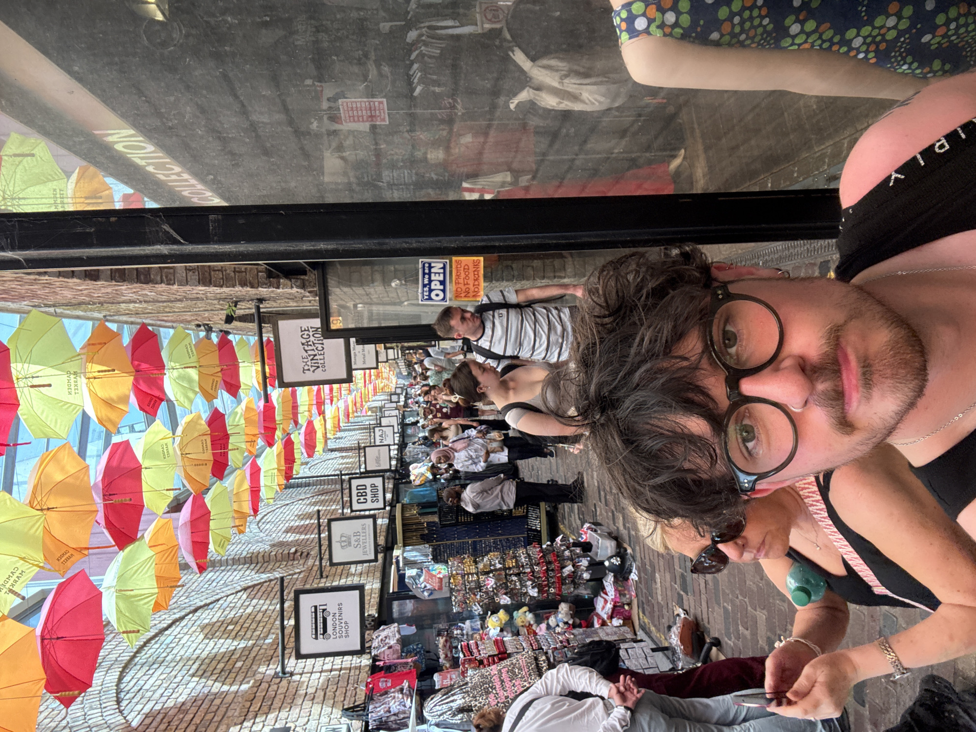 The image shows a selfie of two people, a man and a woman, with a crowded street scene as the backdrop. The man is closer to the camera, wearing glasses and a black top. The woman is beside him, wearing sunglasses and also a black top with a pink strap. They are standing on a brick-paved street. In the background, a row of shops lines the street, with signs indicating businesses such as "LONDON SOUVENIRS SHOP," "S&B JEWELLERS," and "CBD SHOP." Numerous people are walking along the street. Above the street, a canopy of colorful umbrellas creates a vibrant overhead display. A sign that reads “VINTAGE COLLECTION” is visible. A window behind them has a reflection of the scene, including an "OPEN" sign and a person in a striped shirt.