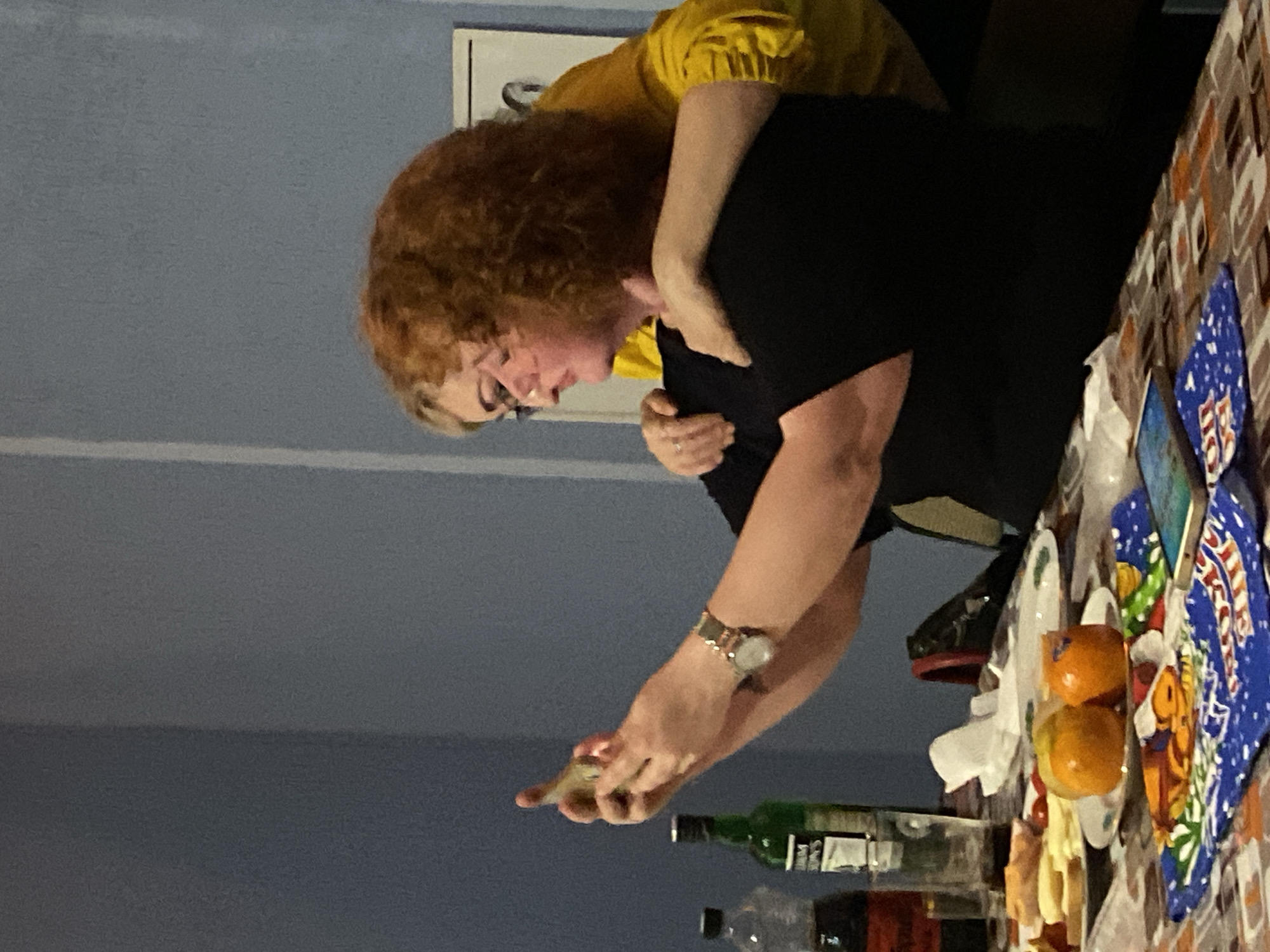 A woman with red curly hair and glasses is taking a picture with a cell phone at a table. She is wearing a black top and a watch on her left wrist. Another person is standing behind her wearing a yellow top with their arms around her. The table is covered with a patterned tablecloth and has plates of food, fruit, and bottles of liquid. The background is a blue wall. The city of Noviy, Russia is not evident in the image.