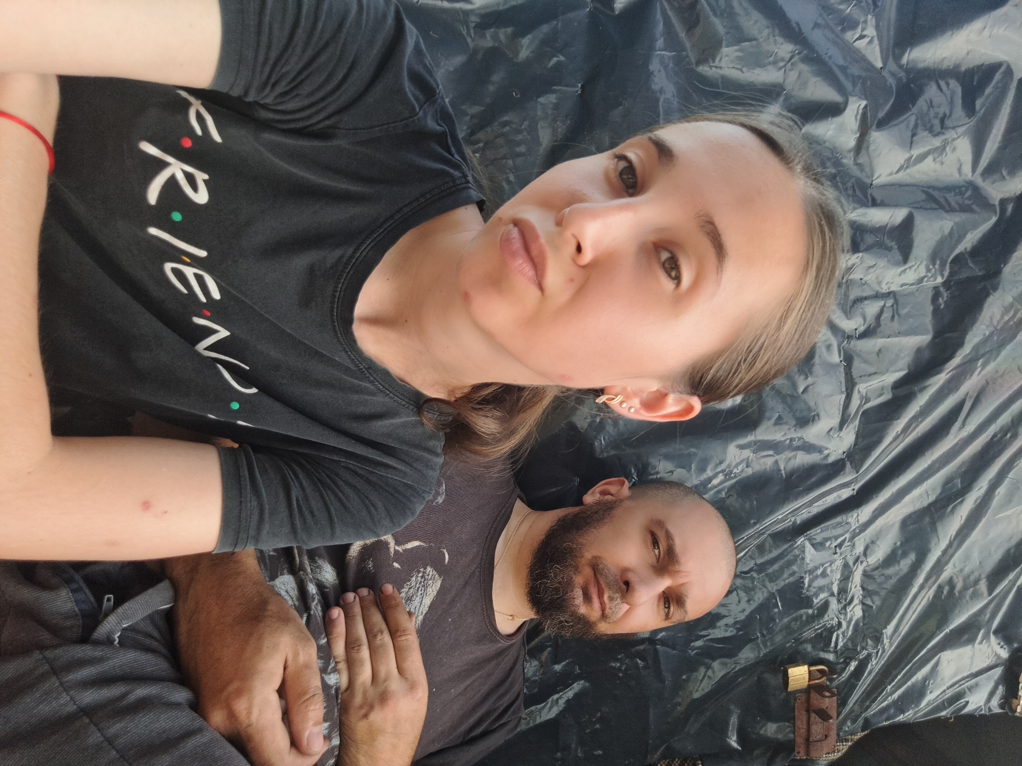 The image shows a young woman and a man sitting side-by-side in front of a dark, crinkled tarp. The woman is in the foreground, facing the camera with a neutral expression. She has light brown hair pulled back and is wearing a black t-shirt with the word "FRIENDS" printed on it in white letters. A red bracelet is visible on her left wrist. To her right, the man has a shaved head and a beard, and is wearing a dark t-shirt with a graphic print. He is looking towards the camera with a serious expression, his right hand resting on his stomach. The tarp behind them has a metallic lock attached to it on the right side. The overall environment appears to be outdoors, possibly in a makeshift shelter or a temporary setup, given the tarp and lock. There are no other prominent objects or individuals visible in the frame. The lighting suggests it is daytime. The scene does not provide specific visual cues to pinpoint the location within Vypasne, Ukraine, beyond the general context of an outdoor setting.