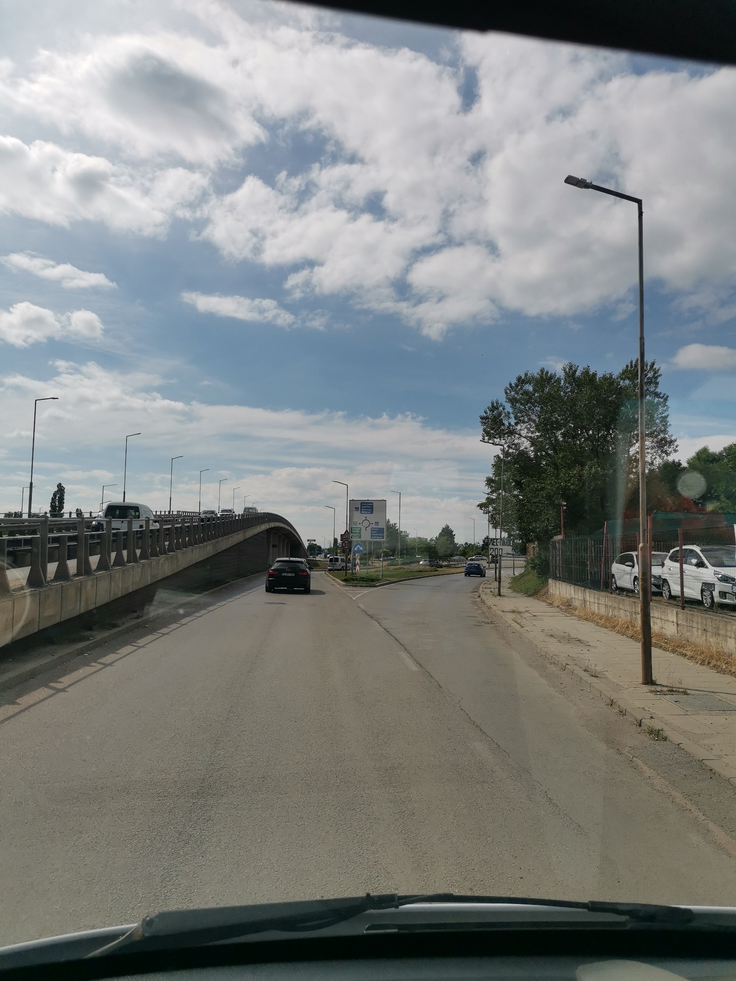 The image shows a view from a vehicle driving on a road in Sofia, Bulgaria. The road curves to the right, and an elevated road or bridge runs parallel to it on the left. Several cars are on both the road and the elevated structure. There are streetlights and trees lining the sides of the road. The sky is partly cloudy, with white clouds against a blue background. There are also traffic signs visible, one of which appears to be a directional sign with text in a Cyrillic alphabet, suggesting the location is in Bulgaria. The overall scene depicts a typical urban road environment.