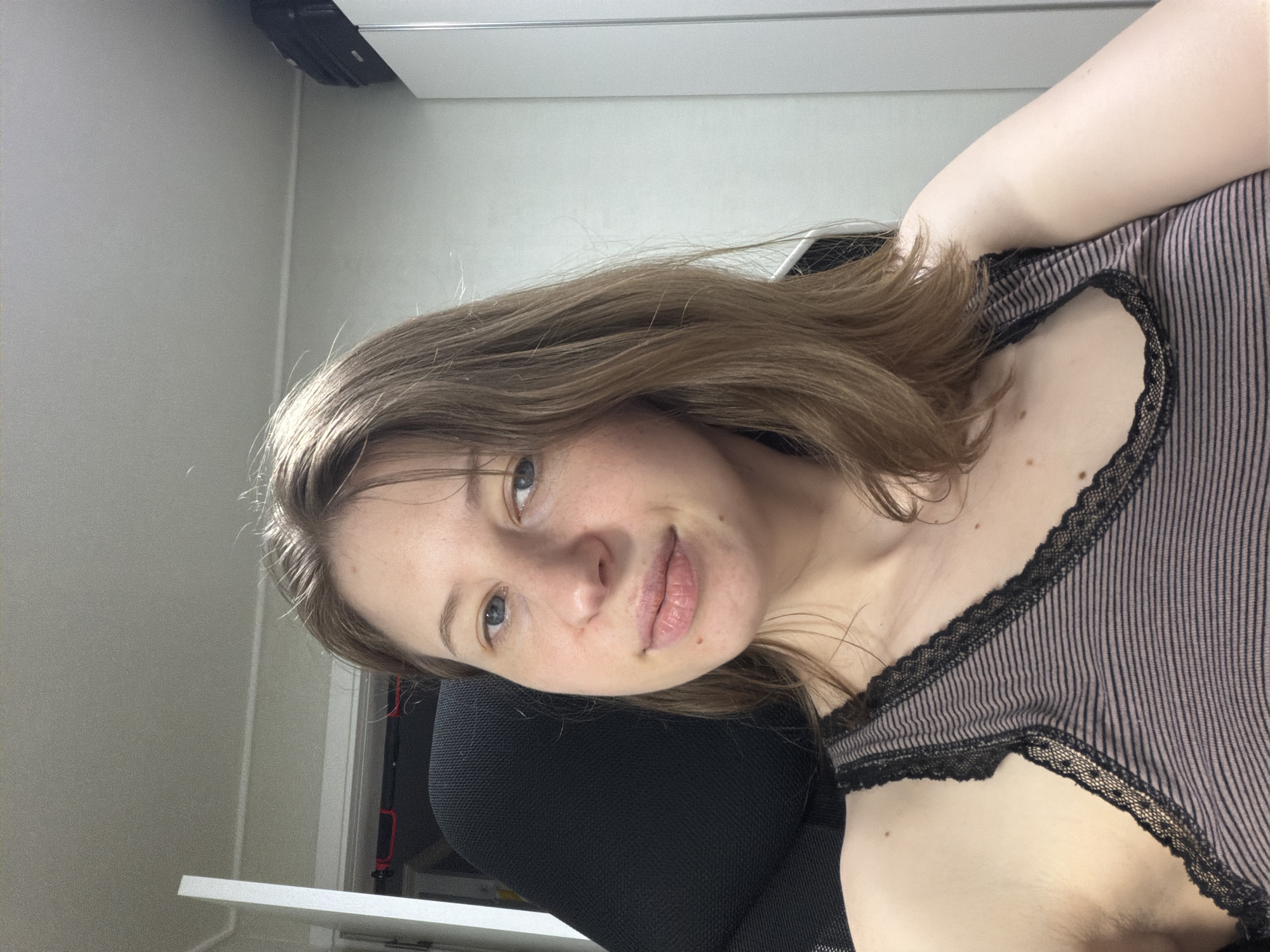 A woman with shoulder-length brown hair and blue eyes is shown in a selfie. She is wearing a striped tank top with lace trim. The woman is seated in a black office chair, and her right shoulder and arm are visible. In the background, there is a white door, a closet, and a wall. The scene appears to be indoors, possibly in a home or office environment. The photograph was taken in Dongducheon-si, South Korea.