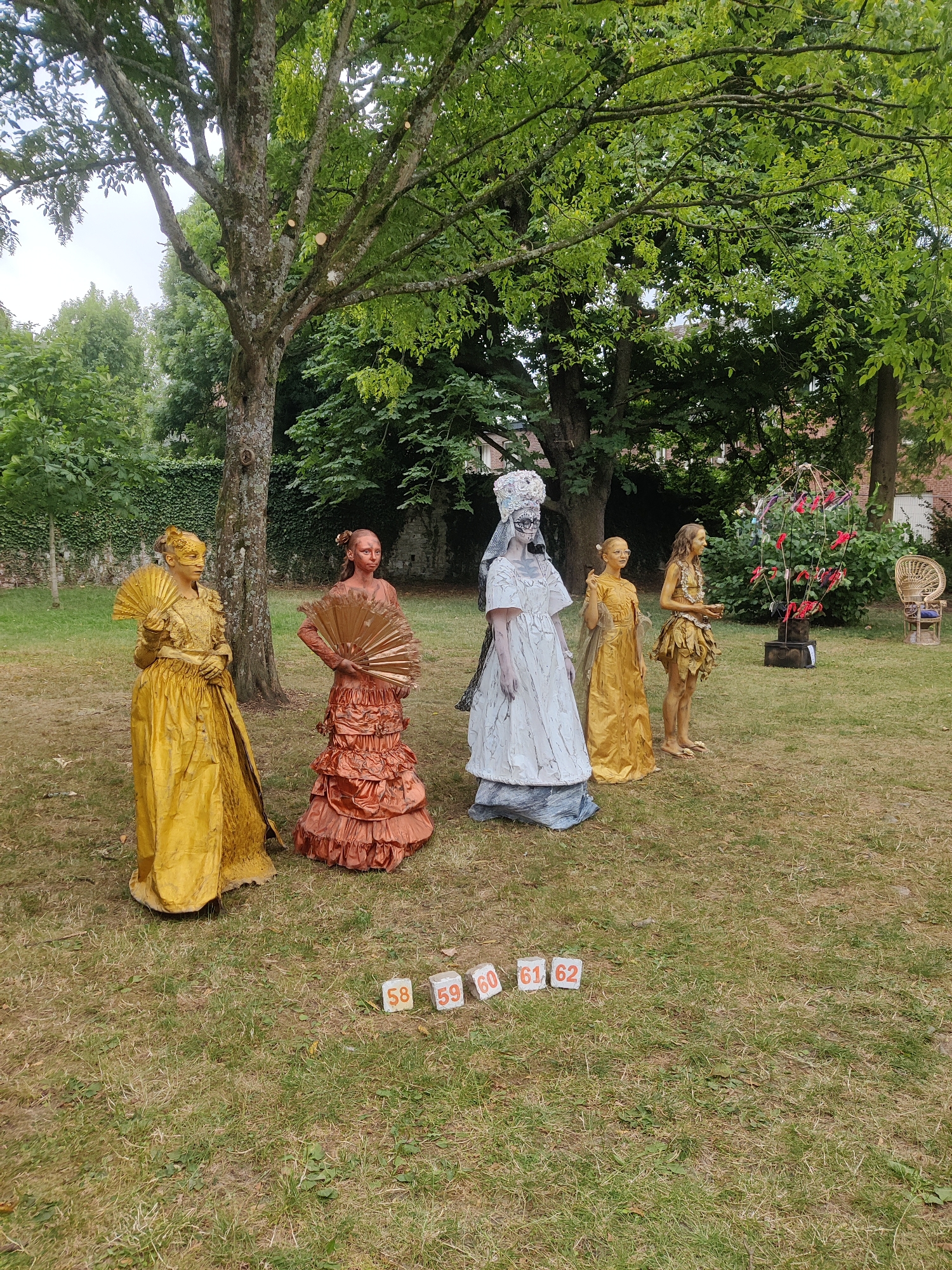 In the city of Marche-en-Famenne, Belgium, a grassy outdoor area is set with five individuals and scattered numbered blocks. In the foreground, from left to right, are five figures painted in various colors and dressed in elaborate costumes. The first figure, on the far left, is painted gold and holds a large, golden fan. The second figure, to its right, is painted a reddish-brown and also holds a similar fan. The third figure is painted white and wears a white headdress and a long, white, layered dress. The fourth figure, to the right of the white-clad figure, is painted gold and wears a long, golden dress. The fifth and final figure, on the far right, is also painted gold and wears a short, leafy-like costume. In front of these figures, the grass is scattered with small, white blocks, each displaying a number: 58, 59, 60, 61, and 62. The background features lush green trees, a stone wall, and in the far right, a wicker chair.
