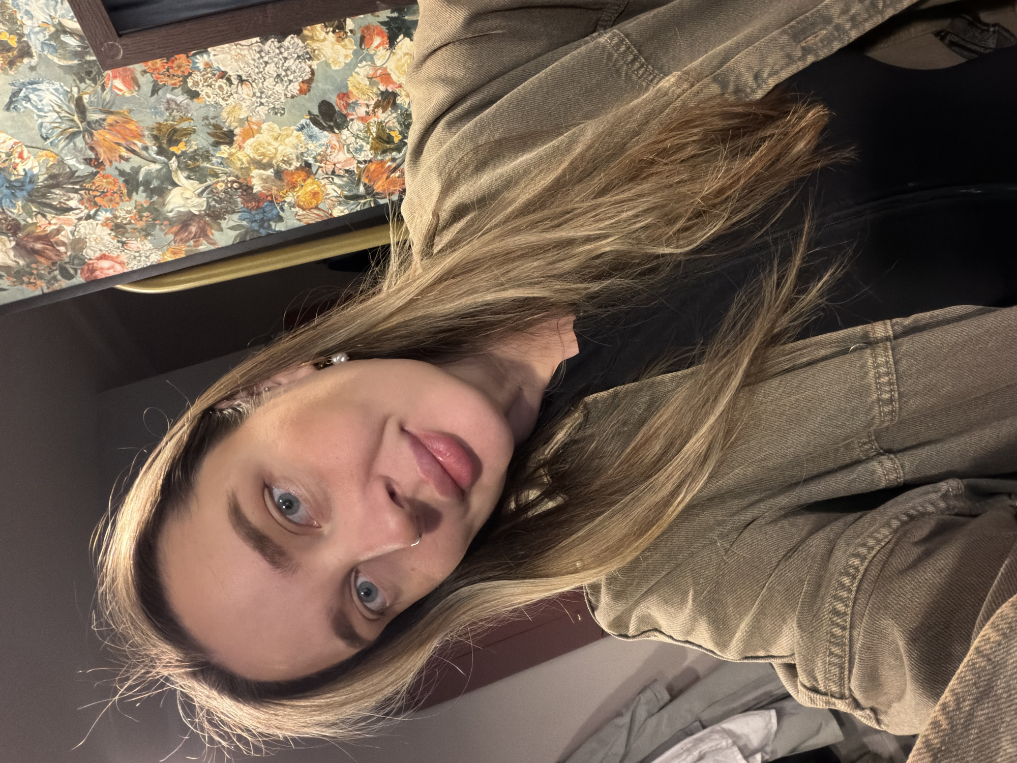 The image displays a young woman with long, straight light brown hair and striking blue eyes, gazing directly at the viewer. Her head is tilted to the side, suggesting the photograph was taken from a lower angle or is rotated. She has a subtle silver hoop nose piercing and a small pearl-like earring. Her lips are slightly parted, conveying a calm and natural expression.
She is dressed in a dark top, likely black, worn beneath a light khaki or olive green denim jacket. The jacket features visible stitching and a textured fabric. The background immediately behind her head showcases a rich, vintage-style floral pattern, possibly on wallpaper or a tapestry. This pattern includes an array of flowers in shades of orange, yellow, blue, white, and green set against a muted blue-green backdrop. Below this pattern, a slender, golden-colored horizontal bar is discernible. The remainder of the background is dark and indistinct, characteristic of an indoor environment. The lighting is soft and appears to be artificial, casting a gentle glow on her face and hair. There is no visible text in the image. The scene is set in Moscow, Russia.