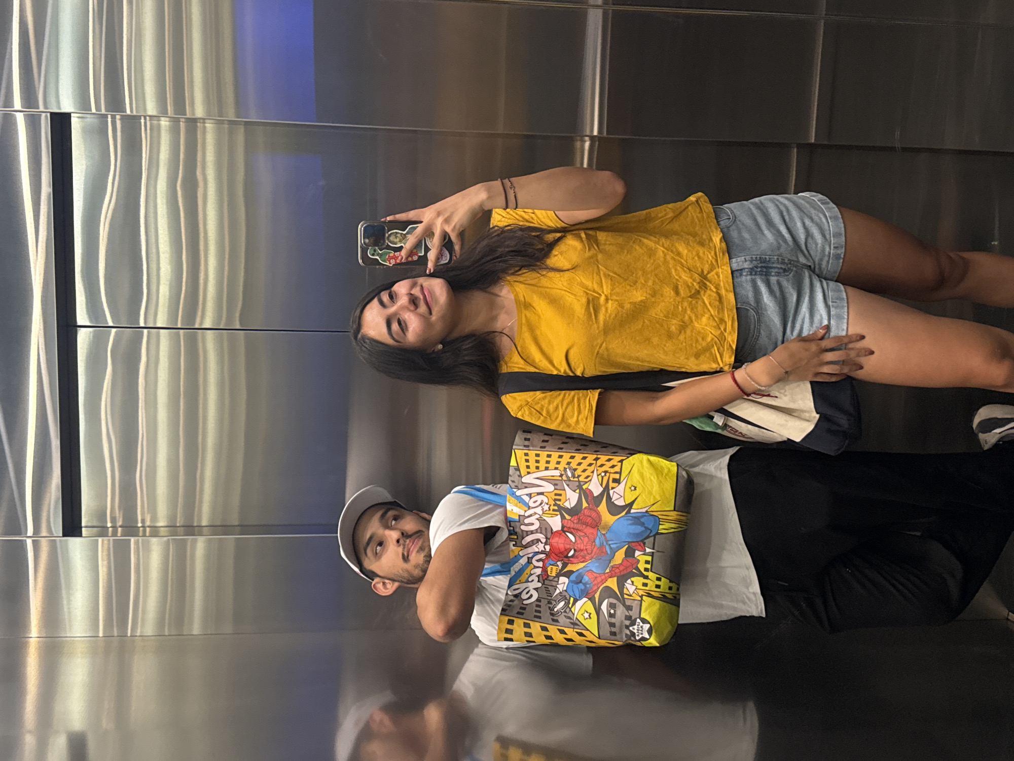 Two individuals are in an elevator, posing for a selfie. The woman is on the right, wearing a yellow t-shirt and denim shorts, holding up a phone with stickers on the case. The man is on the left, wearing a white t-shirt, black pants, and a baseball cap; he is carrying a tote bag with a Spiderman design. The elevator walls are stainless steel. This image was taken in Puebla, Mexico.