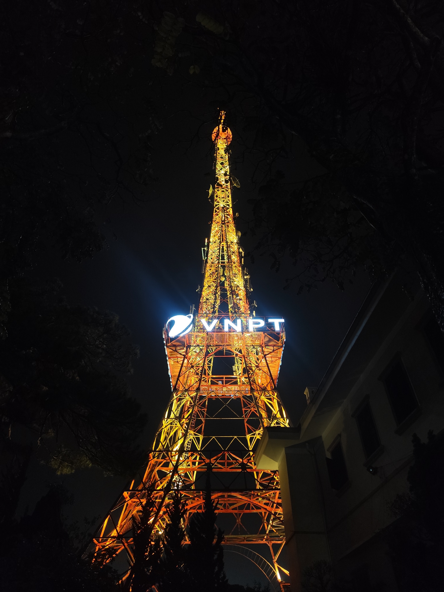 The image presents a striking nighttime view of a tall, brightly illuminated telecommunications tower, likely located in Da Lat, Vietnam. The tower, constructed from a complex lattice of steel girders, glows with warm orange and yellow light, dominating the frame and ascending towards a narrow spire topped with antennas.
Prominently displayed near the upper section of the tower is an illuminated sign featuring a blue and white logo, which includes a stylized globe or "P" shape, followed by the white text "VNPT" (Vietnam Posts and Telecommunications Group). This sign emanates a distinct white light, contrasting with the tower's warmer hues.
The scene is set at night, with the sky appearing dark and clear, though a slight atmospheric haze might be present around the brightest lights. Dark silhouettes of trees and foliage frame the tower on the left and bottom, their branches partially visible against the artificial glow. On the right side of the image, a portion of a building with dark windows and a relatively simple, utilitarian architectural style is visible, mostly in shadow.
No human subjects or specific activities are depicted. The photograph captures the static grandeur of the modern infrastructure against a serene nocturnal backdrop, emphasizing its height and technological presence.
