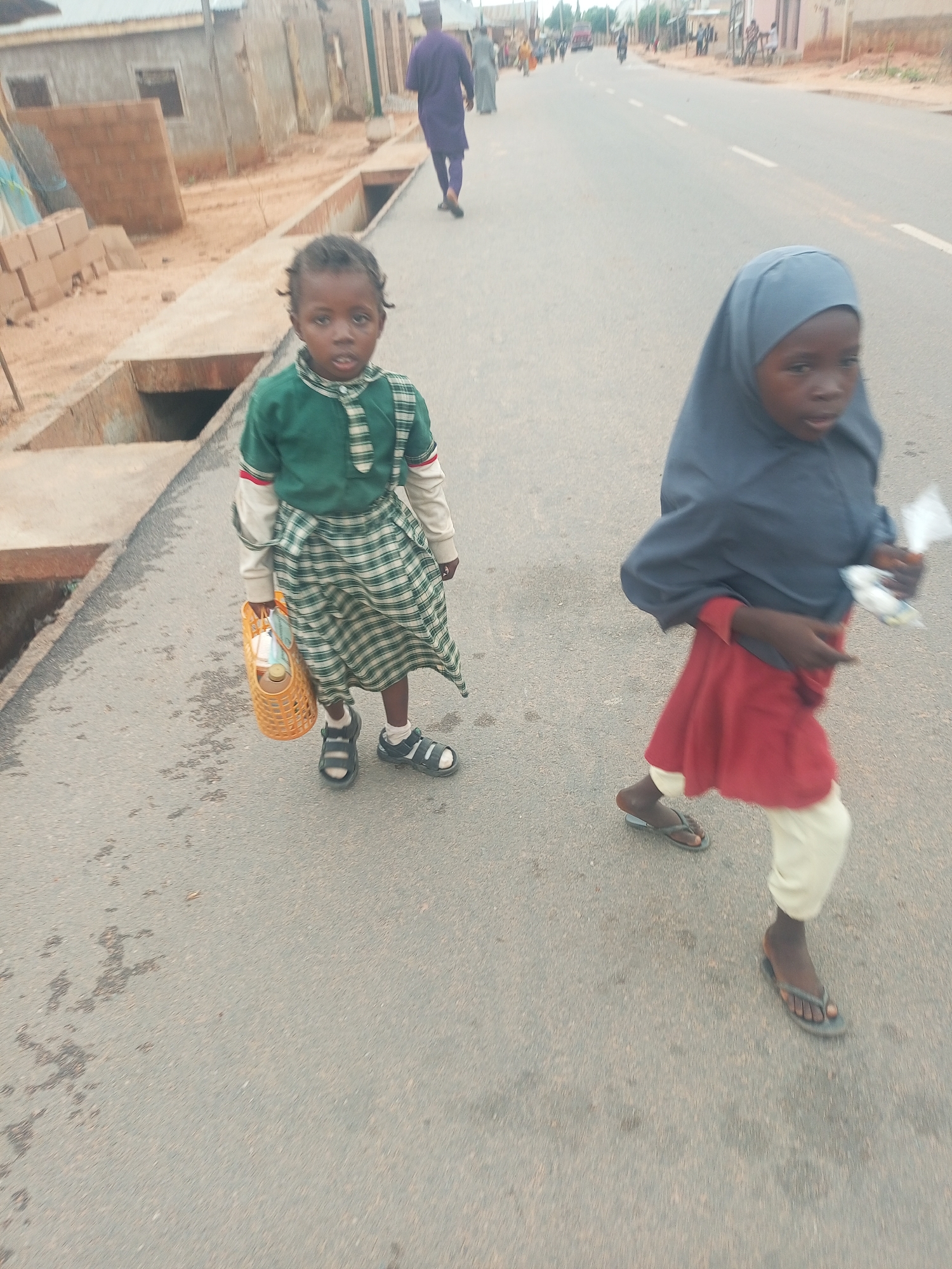 Two young girls are standing and walking on a paved road in Tempure, Nigeria. The girl on the left is standing still, holding a yellow woven basket filled with items. She is wearing a green and white plaid skirt and a green collared shirt with a tied neck. The girl on the right is walking, wearing a grey hijab, a red dress, and light-colored trousers. A few other people are walking further down the road. Some buildings are visible in the background.