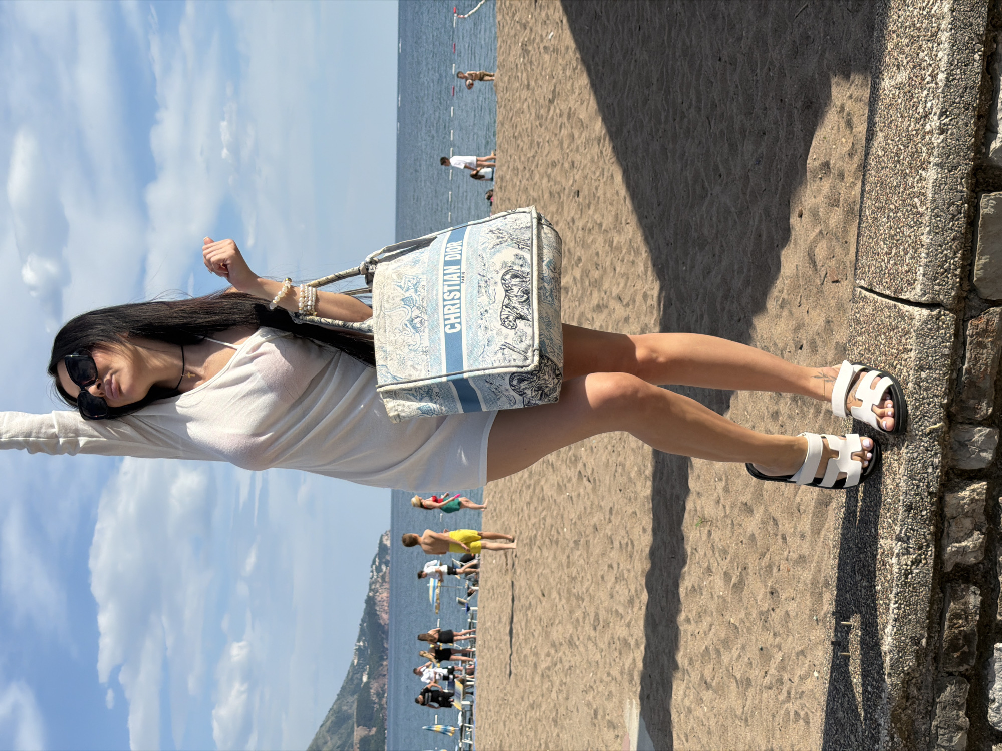 The image shows a woman posing on a beach in Boreti, Montenegro. She is wearing a white dress and sandals, and carrying a white and blue Christian Dior tote bag. She is wearing bracelets on her left wrist and sunglasses. She is standing next to a stone wall that separates the beach from the street. In the background, there are people on the beach, some swimming in the sea. A rocky mountain is visible. The sky is blue with some clouds.