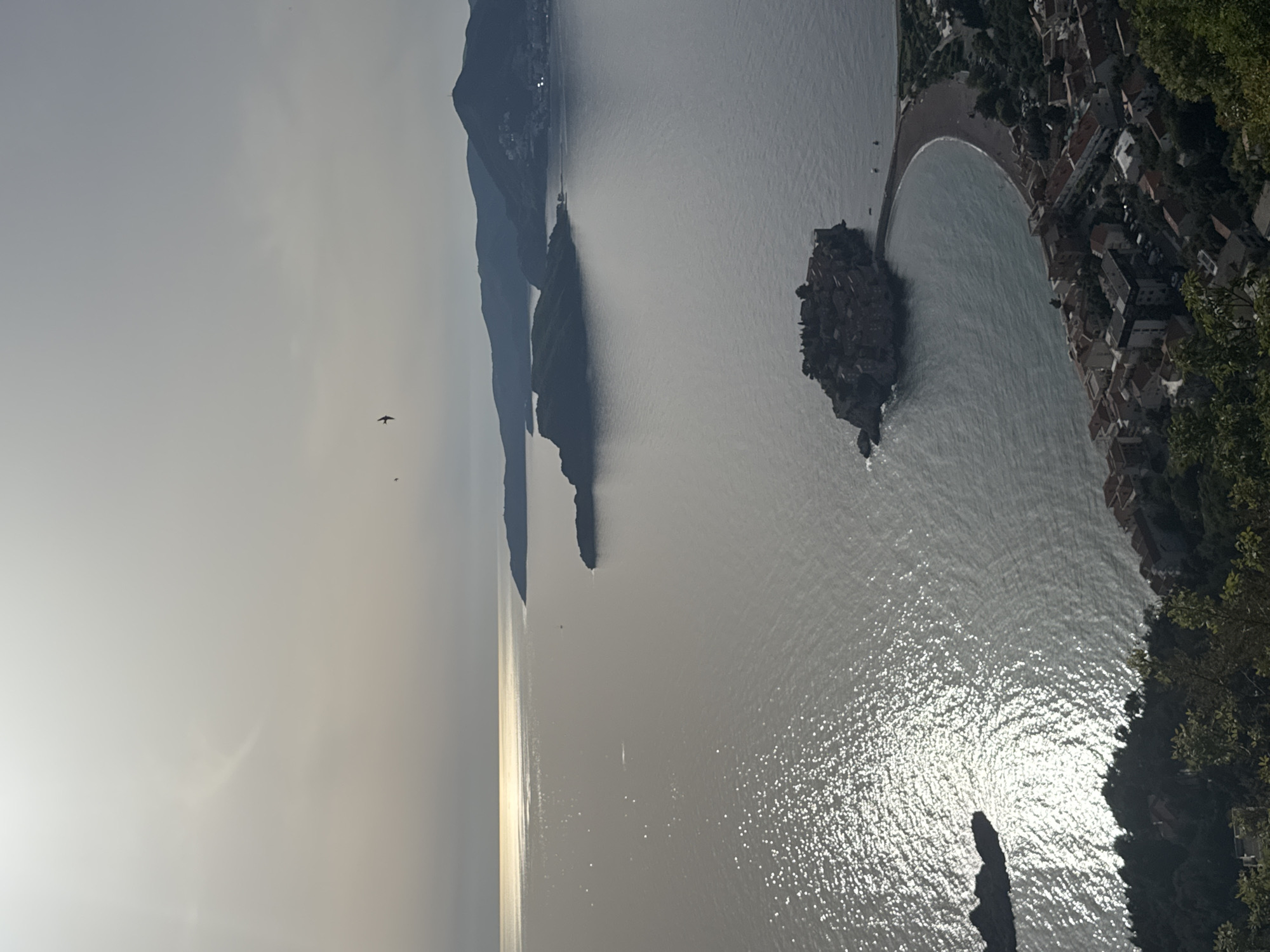 This is an aerial view of Sveti Stefan, Montenegro. The Adriatic Sea is visible, with islands, including Sveti Stefan itself, connected to the mainland by a narrow isthmus. The town's red-roofed buildings line the coastline. The sky is overcast. The image is rotated 90 degrees counterclockwise.