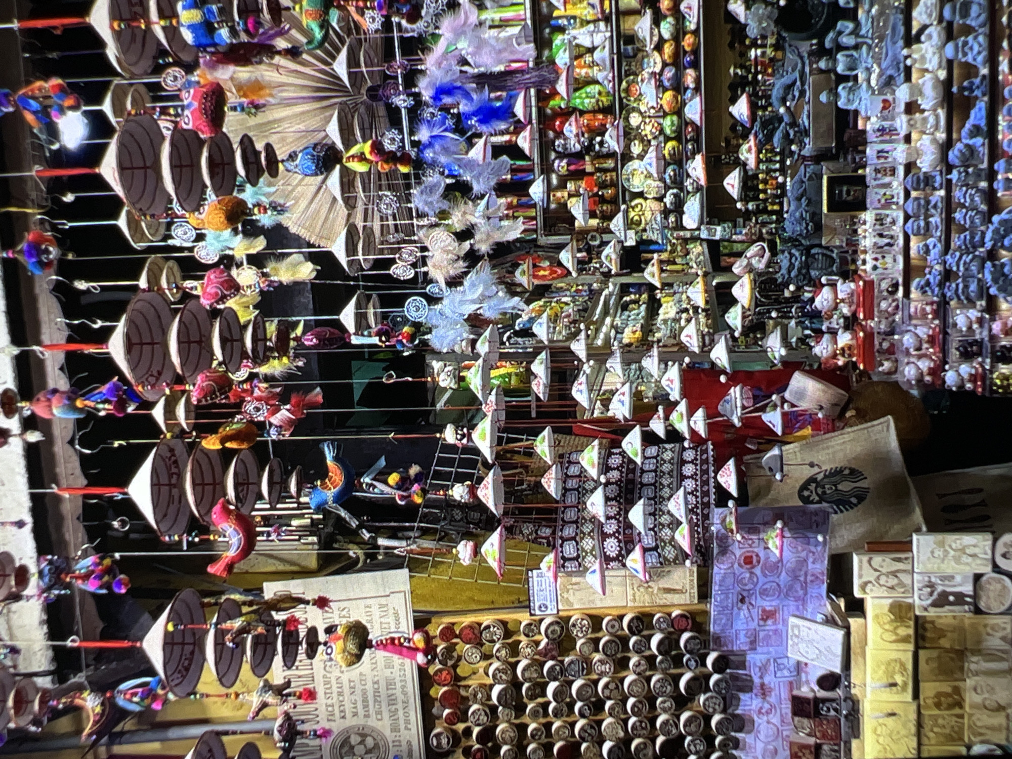 This image presents a vibrant, cluttered display of various handicrafts and souvenirs, likely from a market stall or shop. The scene is densely packed with items, mostly hanging from horizontal lines, and appears to be illuminated by artificial light, suggesting an indoor setting or nighttime market.
The primary subjects are numerous miniature conical hats, reminiscent of traditional Vietnamese "non la." These small hats, often white or light-colored with dark rims and decorative patterns, are strung together with other colorful ornaments such as small fish, birds, and other fanciful shapes, many adorned with soft feathers in shades of blue, pink, and white.
In the background, a vast assortment of smaller items is visible, including what appear to be colorful magnets, pins, or decorative buttons arranged on shelves. Towards the right, displays of carved wooden blocks or stamps are prominent, and a brown bag featuring the Starbucks logo can be seen.
The atmosphere is bustling and rich with cultural artifacts. The visible text on a sign at the bottom left confirms the location and nature of the items. The sign lists services and products such as "FACE STAMP," "KEYCHAIN," "MAG NET," "BAMBOO CLP," and "CHOPTICE NANGRAVE." Most notably, the sign includes ":13: HOANG TAN THU HOI VIET NAM" and a "PHONE:0935261," definitively placing the scene in Vietnam.