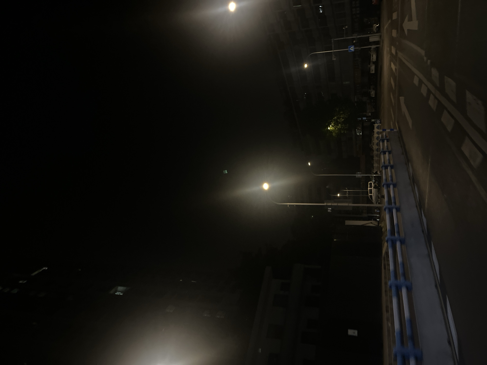 This image captures a nighttime urban streetscape in Yuzhong District, China. The asphalt road in the foreground features white painted markings, including a dashed line and a right-turn arrow. A blue and white barrier runs parallel to the road on the left. The scene is illuminated by several streetlights emitting bright, yellowish-white light, creating prominent lens flare effects in the upper left and right sections of the frame. On the left, multi-story buildings are partially visible, predominantly in shadow, with a few lit windows. On the right, similar multi-story residential structures line the street, displaying balconies and some illuminated interiors. Parked vehicles, including cars and a white van, are visible along the street and in adjacent parking areas. A pedestrian crossing sign (white figure on blue) is affixed to a streetlight pole on the right. A tree with green foliage is centrally illuminated by one of the streetlights. The night sky is dark, and a distinct small green rectangle is observable in the upper central-left portion of the sky. No individuals are visible in the scene.