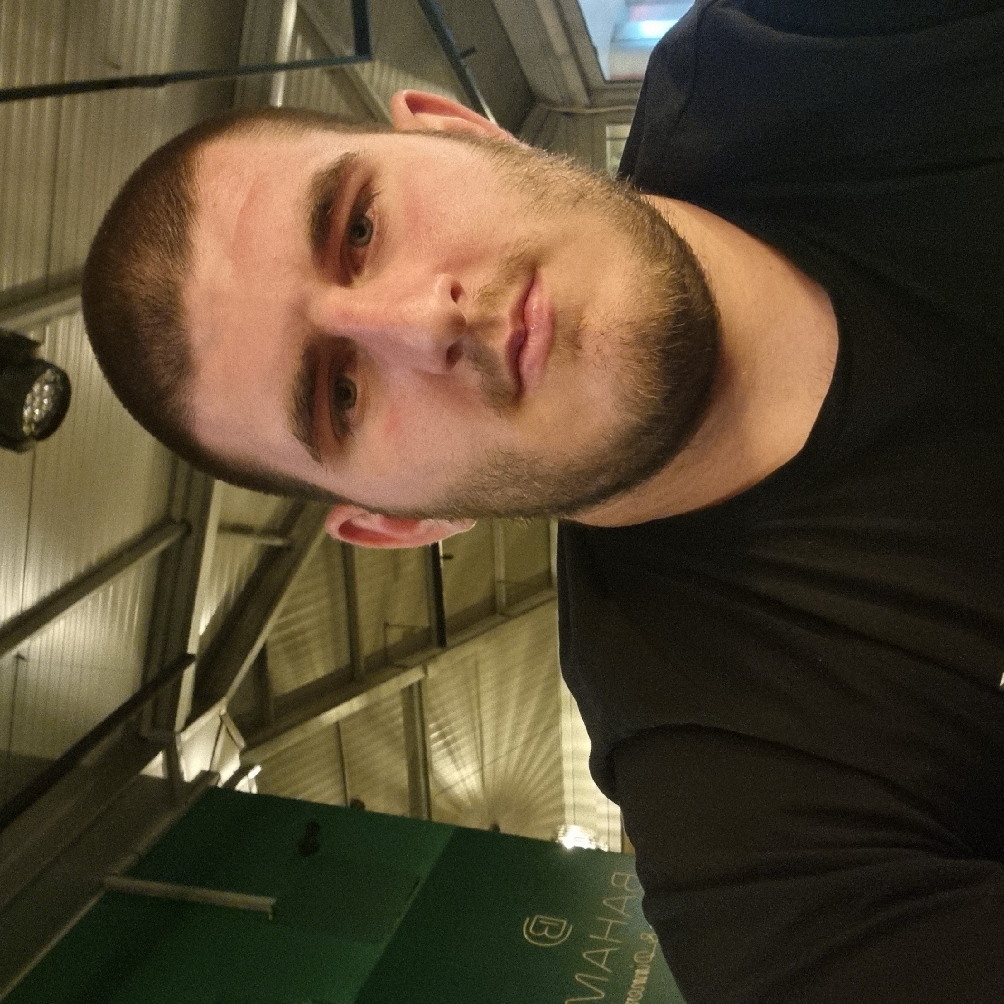 A medium-shot selfie of a fair-skinned male with dark hair cut short, a full beard, and light-colored eyes. He is wearing a black shirt. The background appears to be an indoor space, possibly a restaurant or similar establishment, with industrial-style corrugated metal roofing and exposed beams. A spotlight is visible on the upper left. A green surface with white lettering in reverse order is partially visible in the lower portion of the frame; the lettering includes what seems to be "MAHA" and a logo. No verifiable information is present to concretely verify the presence of the city of Gotse Delchev, Bulgaria.