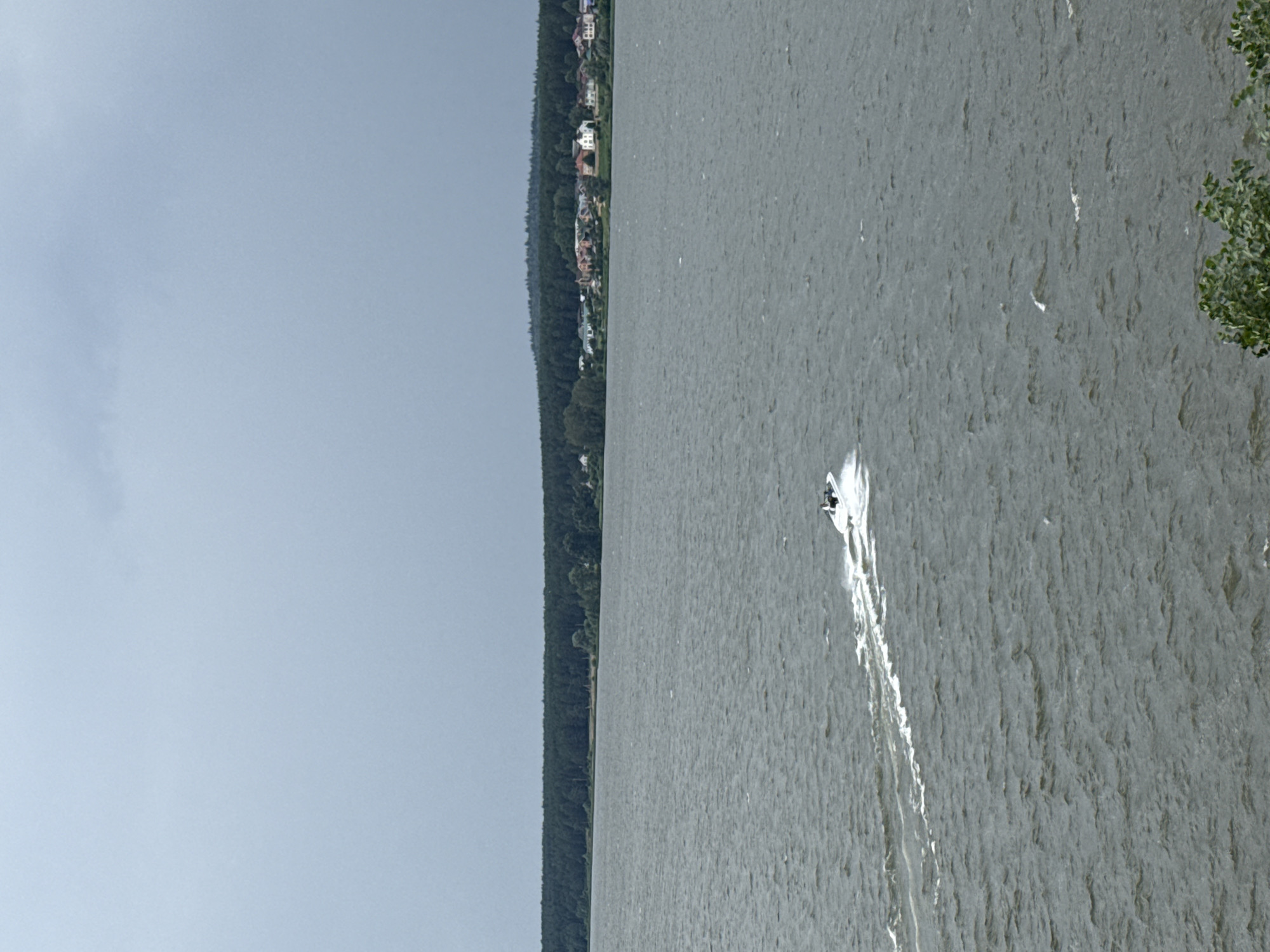 The image depicts a wide, gray, choppy body of water, likely a lake or reservoir, under an overcast sky. In the distance, a dense line of dark green trees forms a forest along the far bank. Behind the forest, a small settlement of houses with red and white roofs is visible. In the middle of the water, a white jet ski is speeding, leaving a white wake behind it. Two figures are visible on the jet ski. The foreground shows the choppy water and the edge of some green foliage at the bottom right. The overall scene suggests a recreational activity on a body of water near a wooded area and a small community.