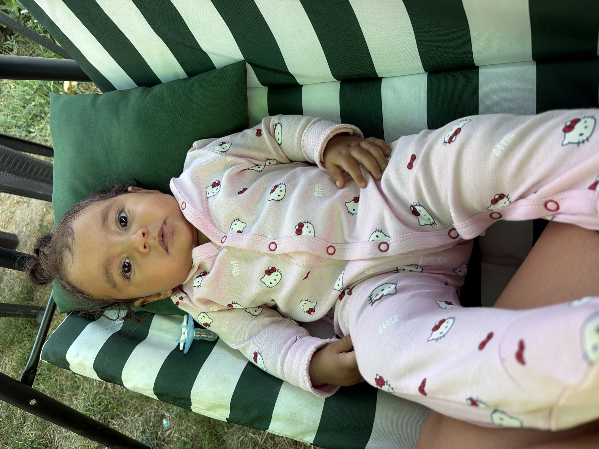 A baby lies on a striped swing seat outdoors. The baby is wearing a pink Hello Kitty pajama set. A green pillow is behind the baby's head. The swing is outdoors, with grass visible in the background. The location is Razhevo Konare, Bulgaria.