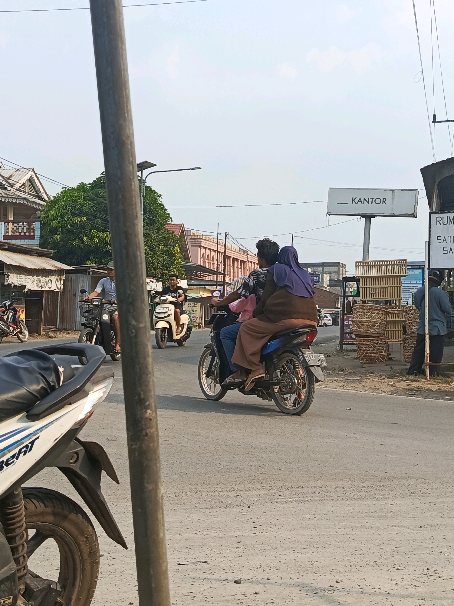 In Megang Sakti I, Indonesia, a street scene unfolds with multiple motorcycles. In the foreground, the rear portion of a white and blue motorcycle with the word "BEAT" visible is parked. A metal pole stands prominently in the middle ground. To the right of the pole, a motorcycle carries two passengers. The rider, wearing a dark t-shirt, is seated in front, while a passenger behind wears a purple headscarf and brown outer garment. Further back, another motorcycle, light beige in color, is ridden by a person in a black t-shirt. On the left side of the image, a man wearing a gray t-shirt and a hat rides a black motorcycle. Buildings line the street, with one having a prominent sign displaying "KANTOR". Several woven baskets are stacked near the side of the road. The sky above is overcast.