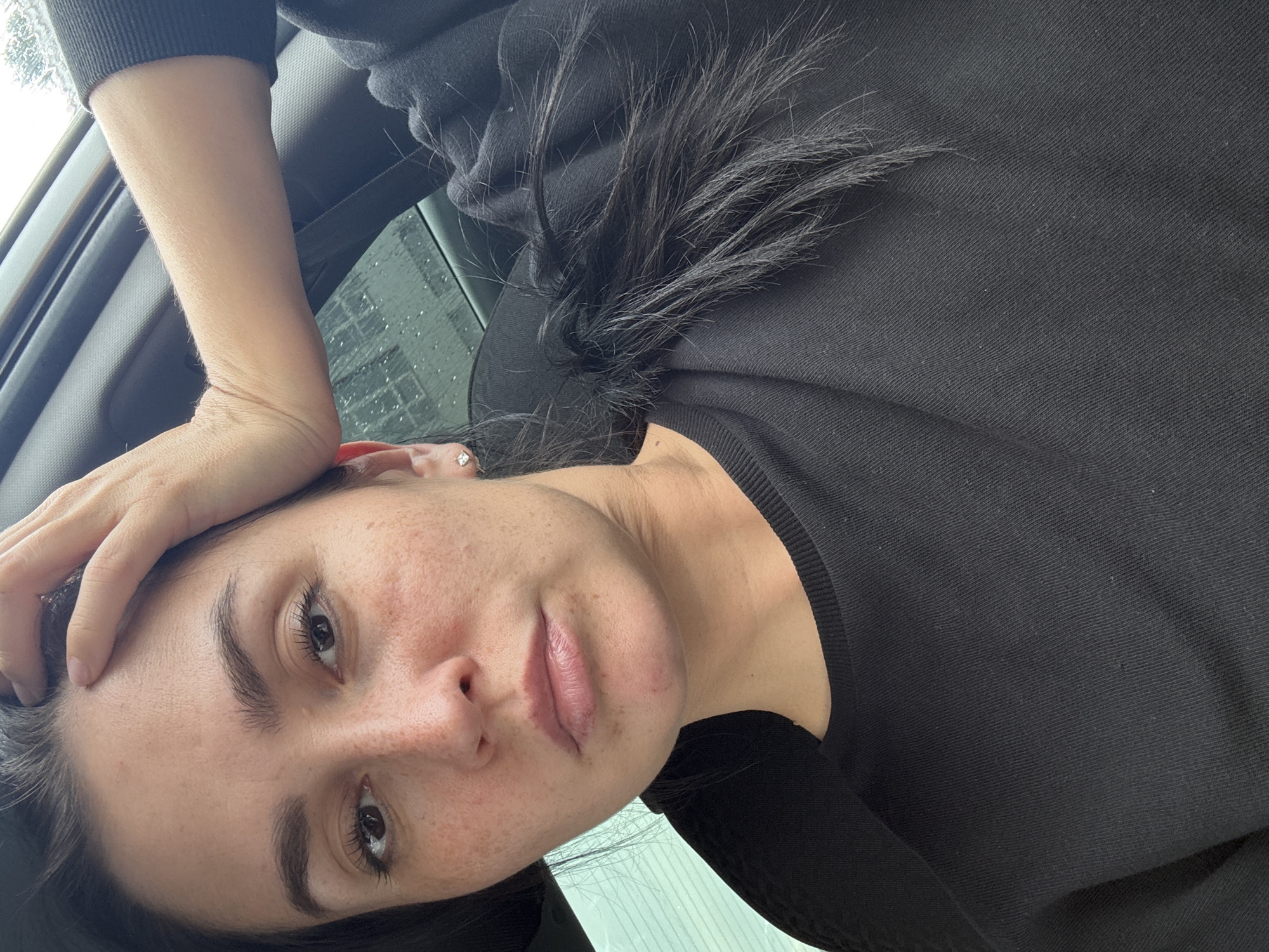 This media file is a close-up, low-angle portrait of a young woman, likely taken outdoors. She is resting her left arm on her forehead, with her hand splayed across her brow. Her eyes are dark brown, and her eyebrows are well-defined. She has a light complexion with visible freckles across her cheeks and nose. Her lips are full and appear to be slightly parted. She is wearing a black long-sleeved top. Her dark hair is loose and falls around her face.
The background suggests an outdoor setting, possibly inside a vehicle, with a window visible to the left. The window appears to have raindrops on it, indicating it might be raining or has recently rained. The lighting suggests it is daytime, but the weather might be overcast. The overall mood of the image is contemplative, perhaps a moment of reflection or fatigue. There is no visible text in the image. The provided location context is Verkhneyarkeyevo, Russia.