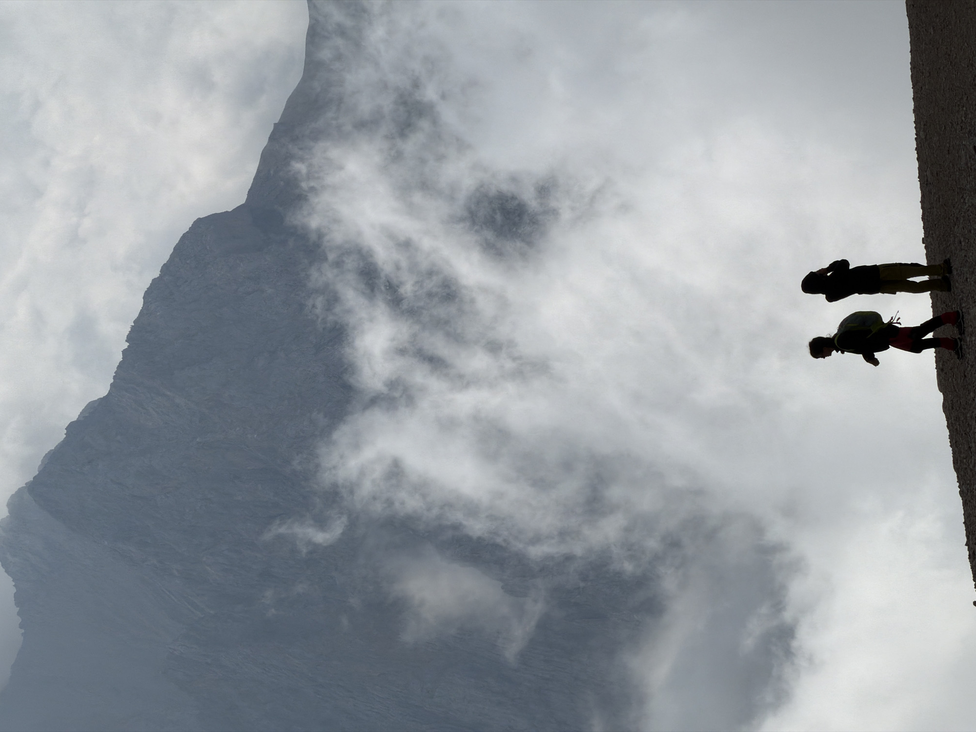 The image depicts two hikers silhouetted against a backdrop of a massive, rugged mountain and swirling clouds. The mountain, which appears to be part of the Wildermieming area in Austria, dominates the upper two-thirds of the frame with its dark, textured slopes. Clouds are obscuring portions of the mountain and are also present in the foreground, creating a dramatic, atmospheric effect. The two figures, a person on the left and another on the right, are walking on a gravelly surface in the lower portion of the image. The person on the left wears a backpack and red leggings, while the person on the right is wearing a hooded jacket and khaki pants. They appear to be walking in the same direction, with the person on the left slightly ahead. The lighting suggests it is daytime, but the overcast conditions create a muted color palette with predominantly grays and dark tones.