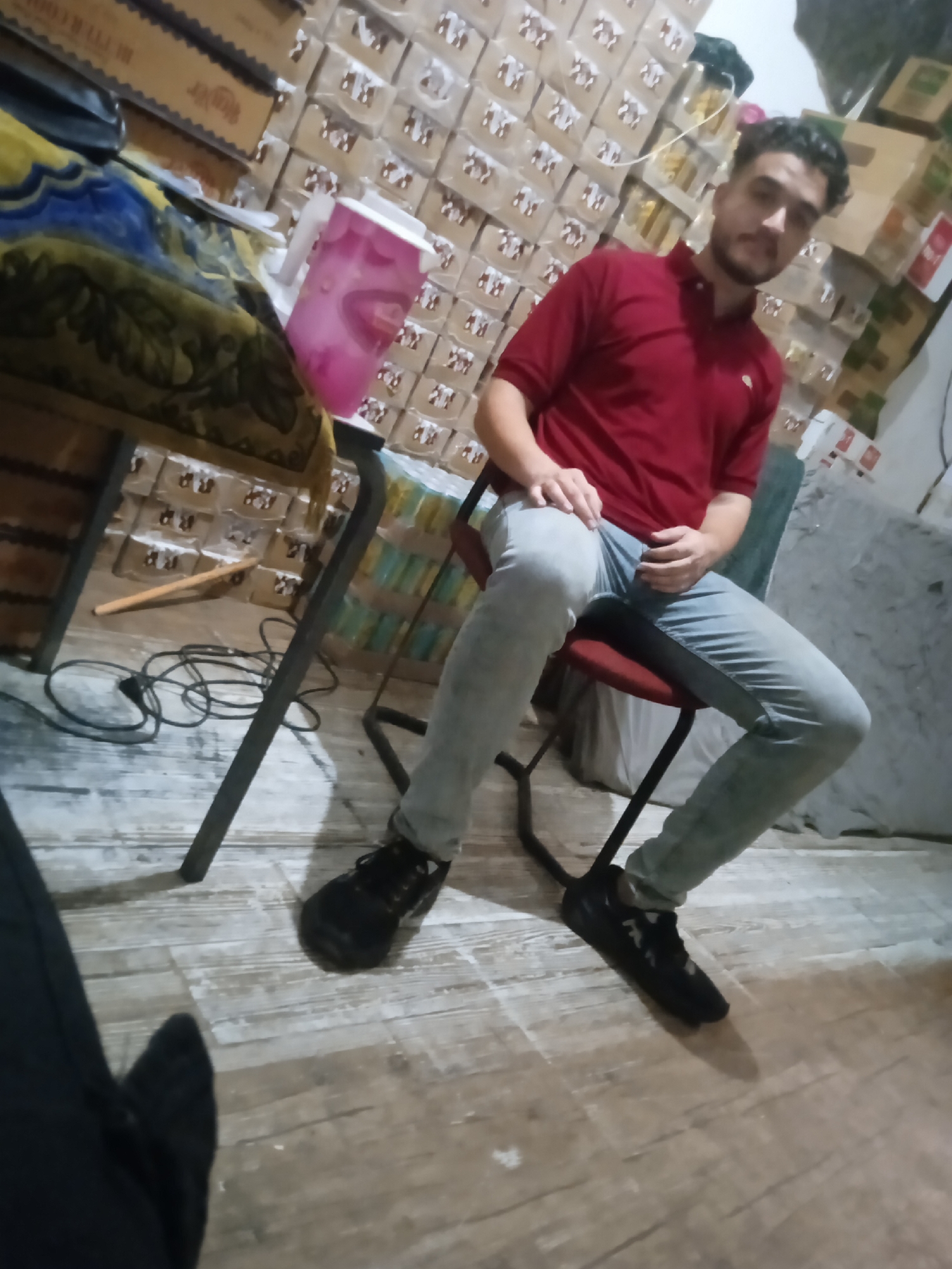 In the city of Gaza, Palestinian Territories, a young man with dark curly hair and a beard, wearing a red polo shirt and light blue jeans, sits on a red chair. He is positioned near a table covered with a yellow and blue patterned cloth. Behind him, stacks of boxes are visible, suggesting a storage area or a shop. A pink pitcher with white lid and handle is on the table, to the left of the man. A black cord snakes across the wooden floor near the chair. The overall scene appears to be indoors, in a dimly lit environment.