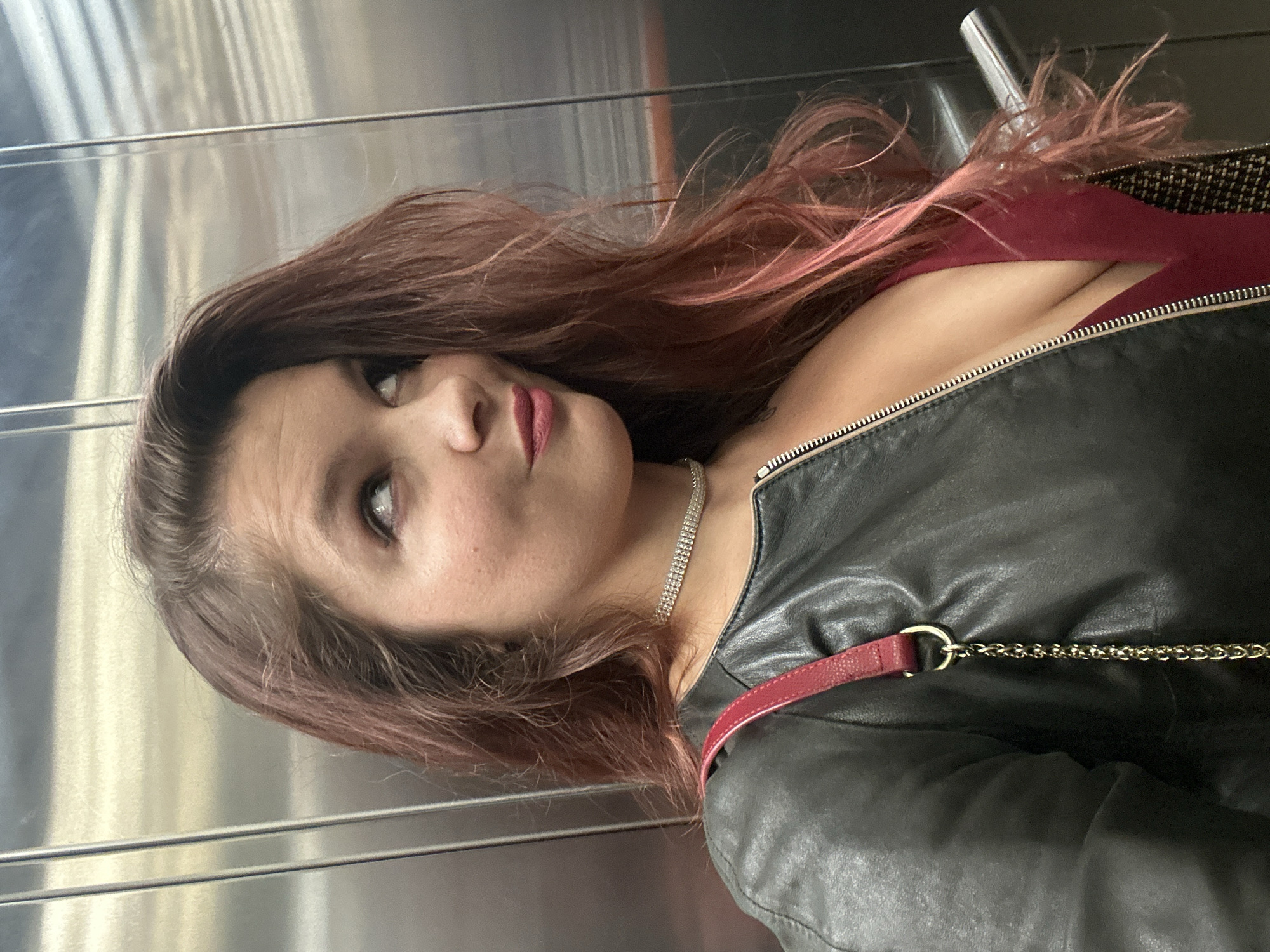 A woman is depicted indoors, likely in an elevator, as indicated by the stainless steel interior. She is fair-skinned with shoulder-length brown hair that has pink highlights. She is wearing a black leather jacket, a red garment underneath, and a silver rhinestone necklace. A red purse strap with a gold chain is visible across her body. She is looking upwards and to the left. The location, Vladimir, Russia, cannot be confirmed from the visual information.