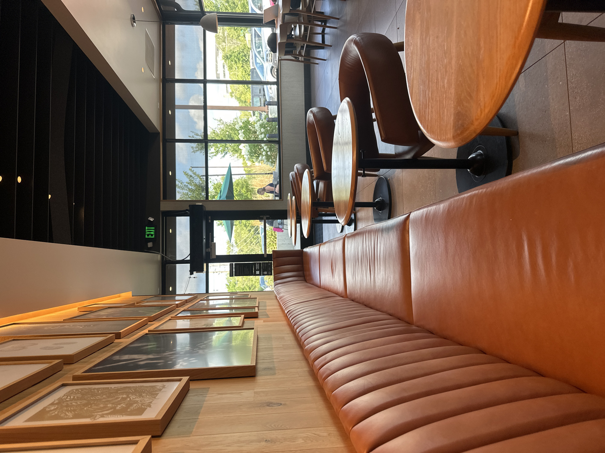 This image depicts the interior of a Starbucks coffee shop in McHenry, United States of America. The scene is oriented towards the right side of the establishment, showcasing a long, ribbed, burnt orange leather booth that extends along a light brown wood-paneled wall adorned with framed artwork. To the right of the booth, several round, light wood-topped tables with black bases are visible, each paired with a brown leather armchair. The background features a large window offering a view of the exterior, with trees, parked cars, and a glimpse of buildings under a cloudy sky. An "EXIT" sign is illuminated green above a doorway to the left of the window. The flooring is a grey, speckled tile. The lighting appears to be a mix of overhead recessed lights and the natural light filtering through the window, with a pendant light hanging near the window. No individuals are clearly depicted in the foreground, though a person can be seen seated at a table in the far background near the window.