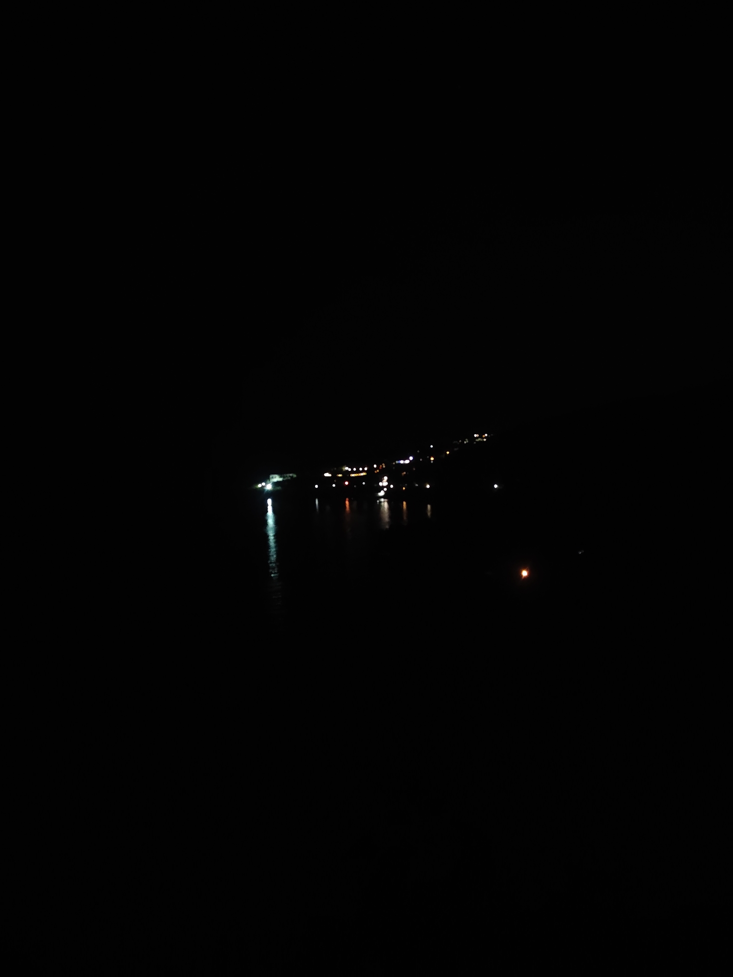 The image shows a dark night scene. A body of water, possibly a lake or bay, occupies the lower portion of the frame. Across the water, in the distance, is a stretch of lights that likely delineate a shoreline with buildings. There are some light reflections on the water's surface. The sky above is entirely black.
