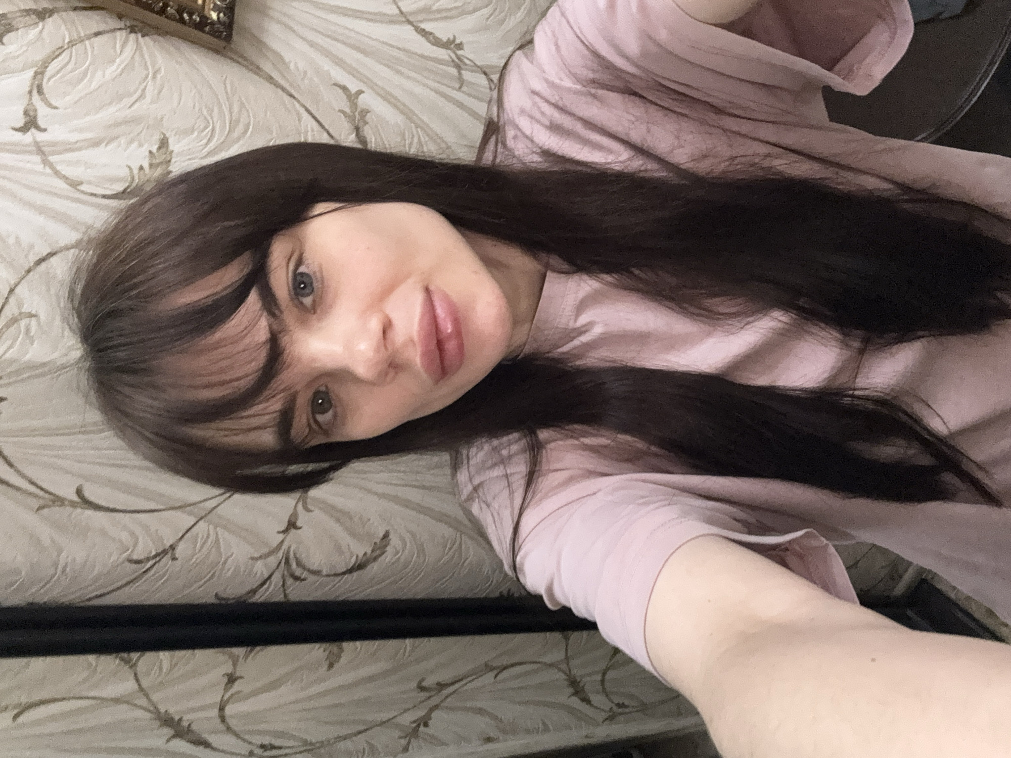 A woman with long dark hair and bangs is lying on a bed with a light-colored patterned bedspread. She is wearing a pink t-shirt and is looking at the camera. Part of a dark-colored metal bed frame is visible along the bottom of the image. The location is not discernible from the image, and a specific location in Rostov-on-Don, Russia, cannot be determined.