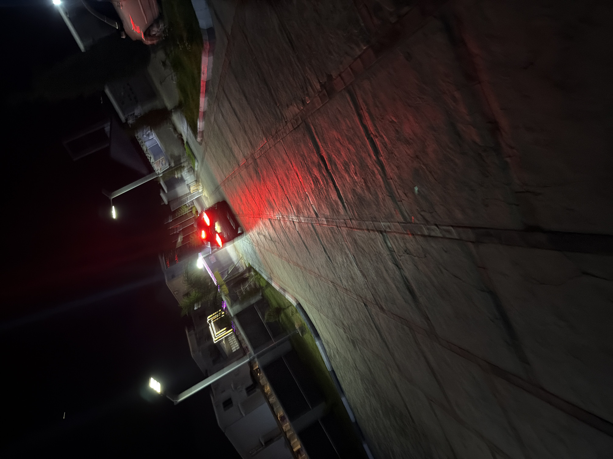 In Bukuru, Nigeria, a car with illuminated red taillights is parked or driving on a paved road at night. The road surface appears to be made of interlocking pavers with distinct lines. To the left of the road, a modern building with white walls and dark windows is visible. Adjacent to the building, there's a garden area with green grass and some plants. Further down the road, a street lamp with a bright white light is illuminating the scene. Another car, possibly a silver SUV, is parked on the right side of the road, partially visible. The overall environment suggests a residential or commercial area illuminated by artificial lights in the darkness.