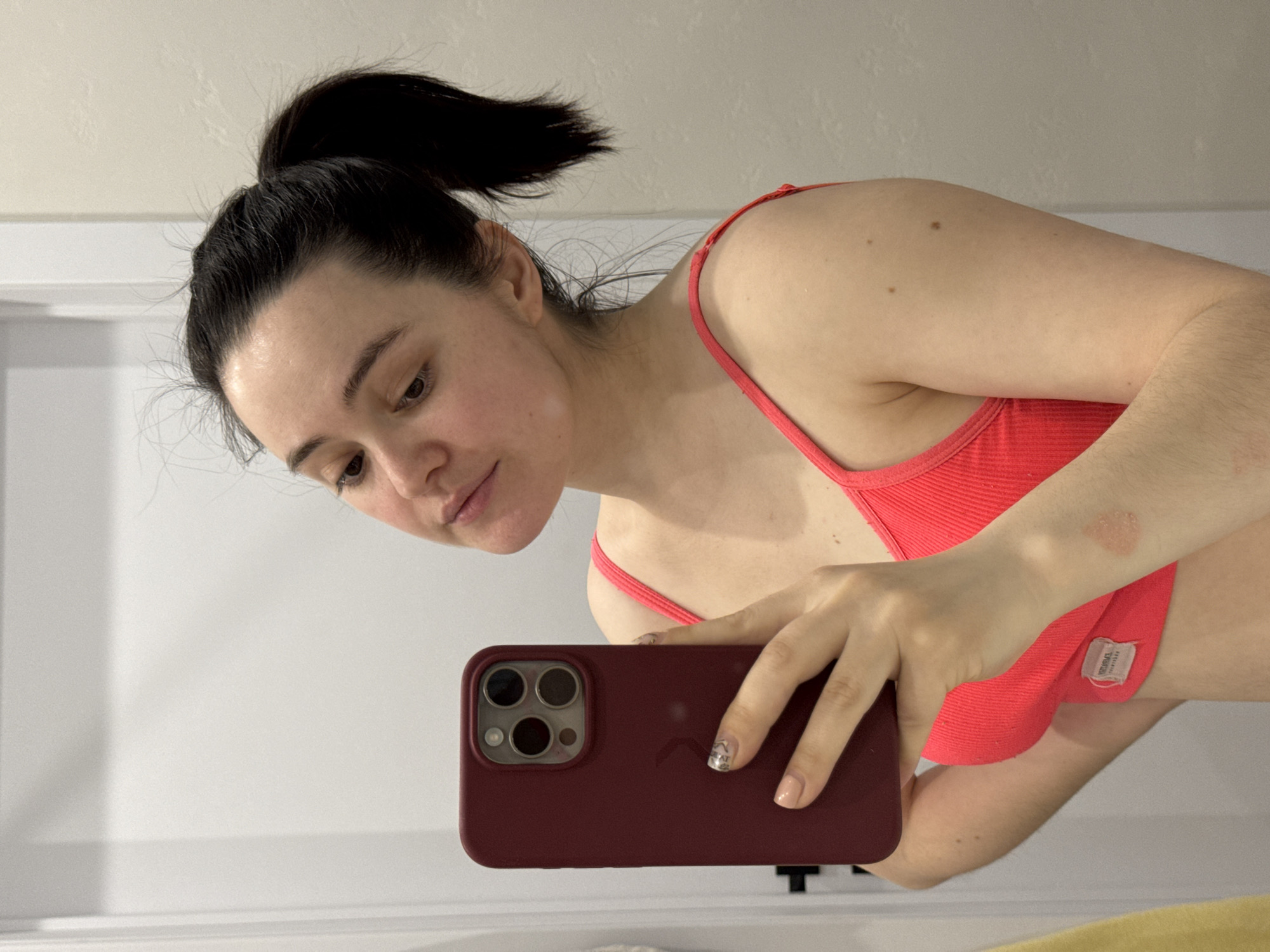 A fair-skinned woman with dark hair pulled into a high ponytail takes a selfie in a bathroom. She wears a red ribbed tank top. Her right arm has a raised, discolored patch of skin. She holds a maroon iPhone with its triple-lens camera facing towards herself. The bathroom has white walls and a white countertop with a mirror. No evidence suggests the location is Izhevsk, Russia.